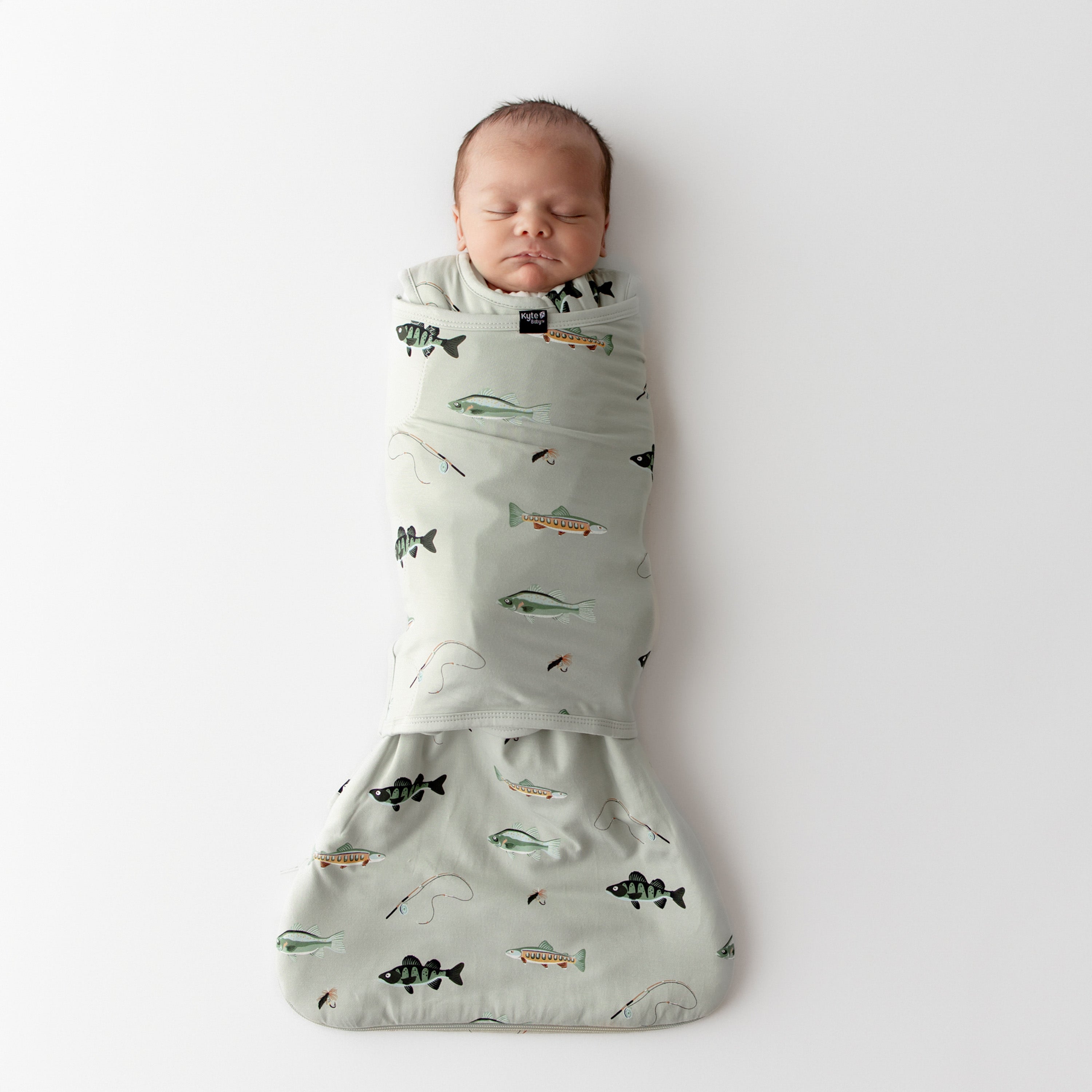 Newborn baby swaddled in a green sleep bag swaddler with fishing prints on a white background