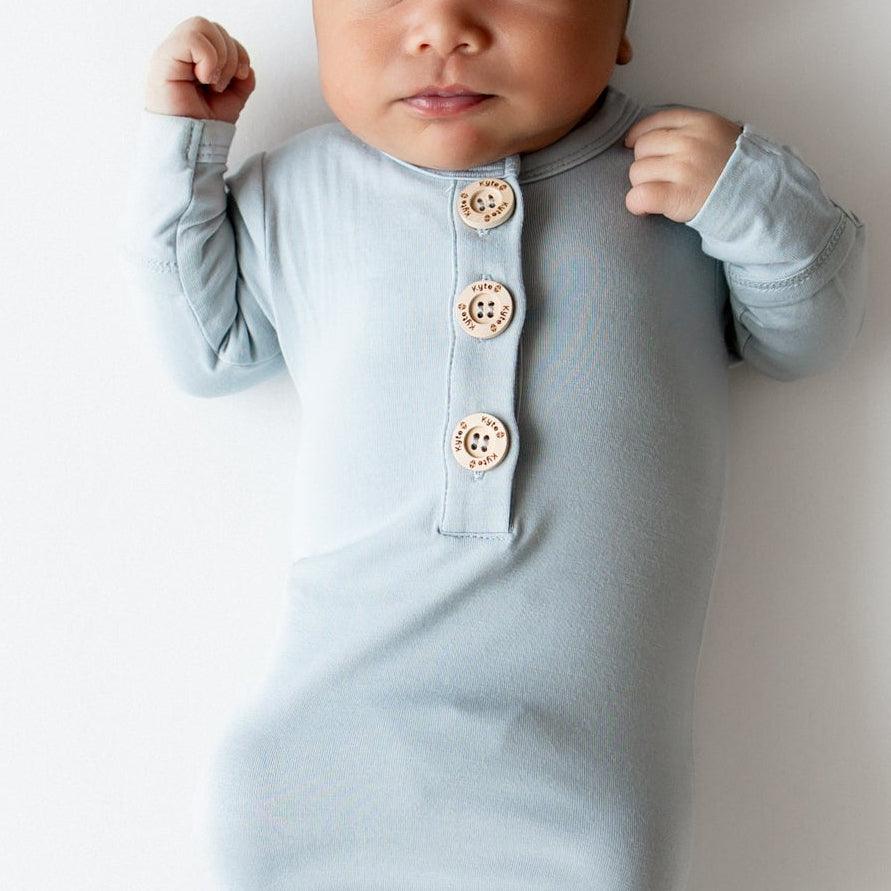 Baby wearing Kyte Baby knotted gown and hat set in a dusty blue color on a white backdrop