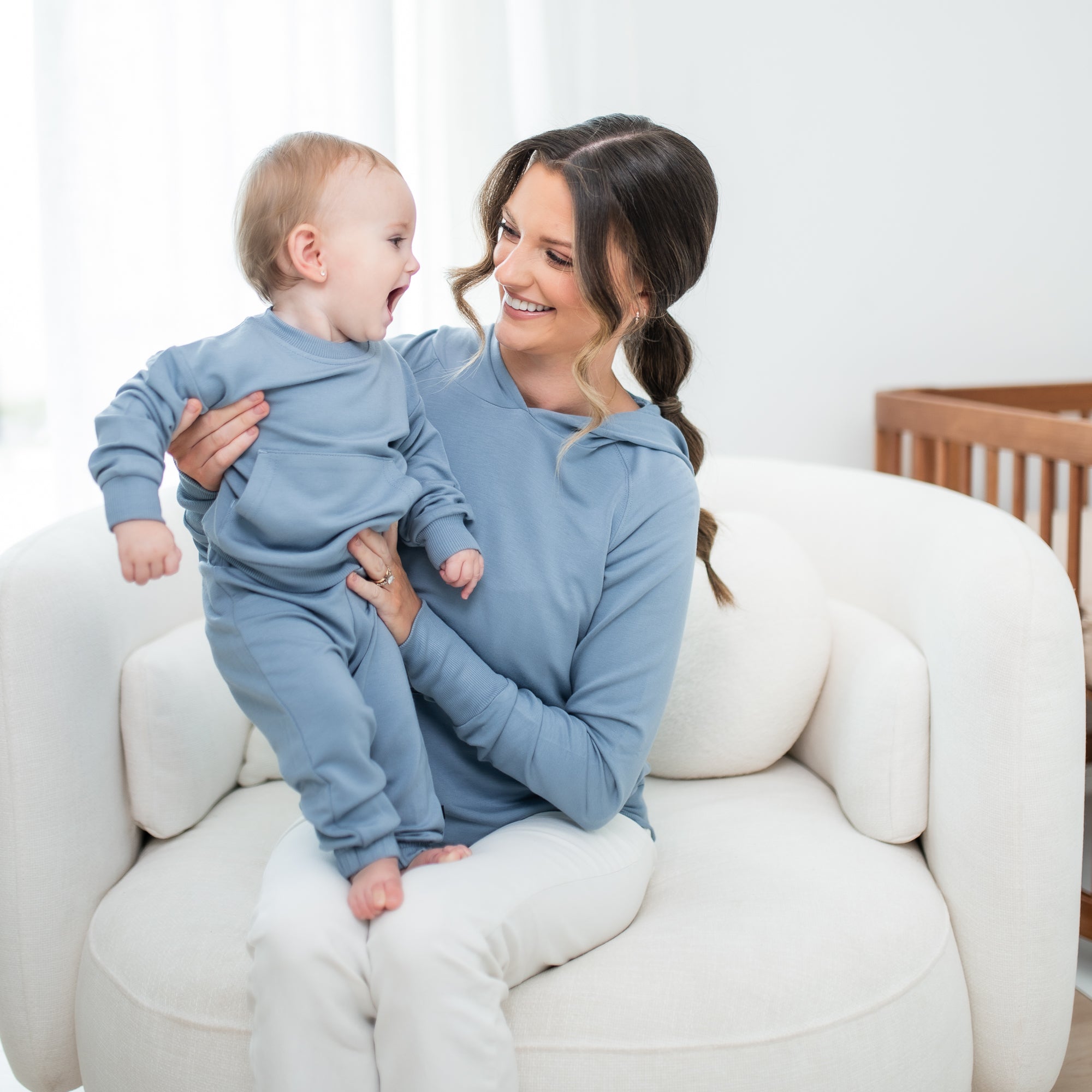 Mother wearing the Women's French Terry Nursing Hoodie in Slate holding her infant ho is in a matching french terry jogger set sitting on a cream colored sofa chair