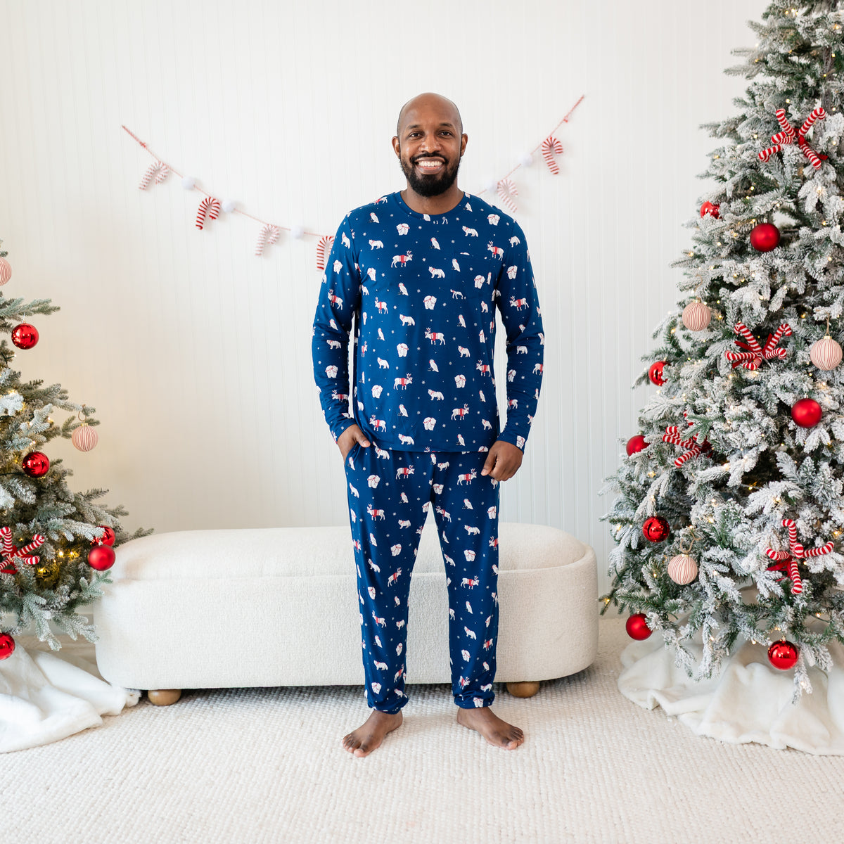 Smiling male model standing in front of a cream ottoman between two decorated Christmas trees wearing the Men's Jogger Set in Frosty Friends