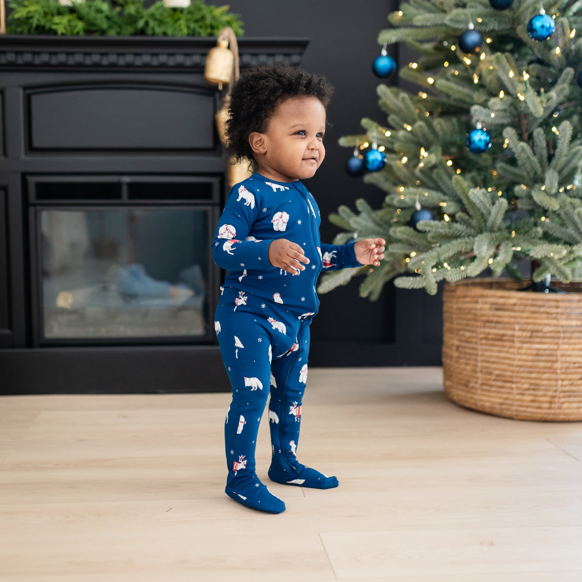Young toddler walking in front of a black fireplace and decorated Christmas tree wearing the Zippered Footie in Frosty Friends