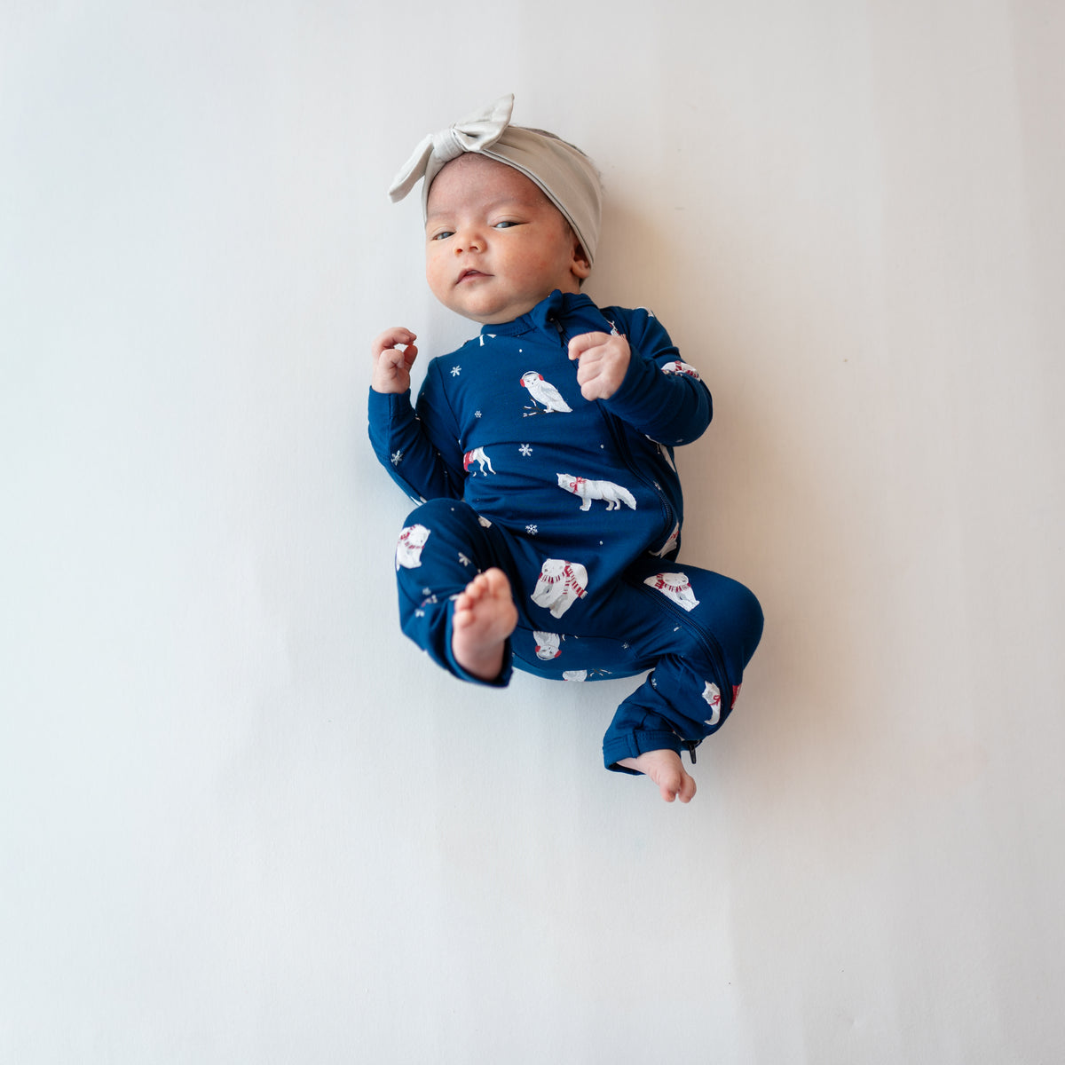 Young infant girl laying down on a light neutral background wearing the Zippered Romper in Frosty Friends with a neutral beige bow