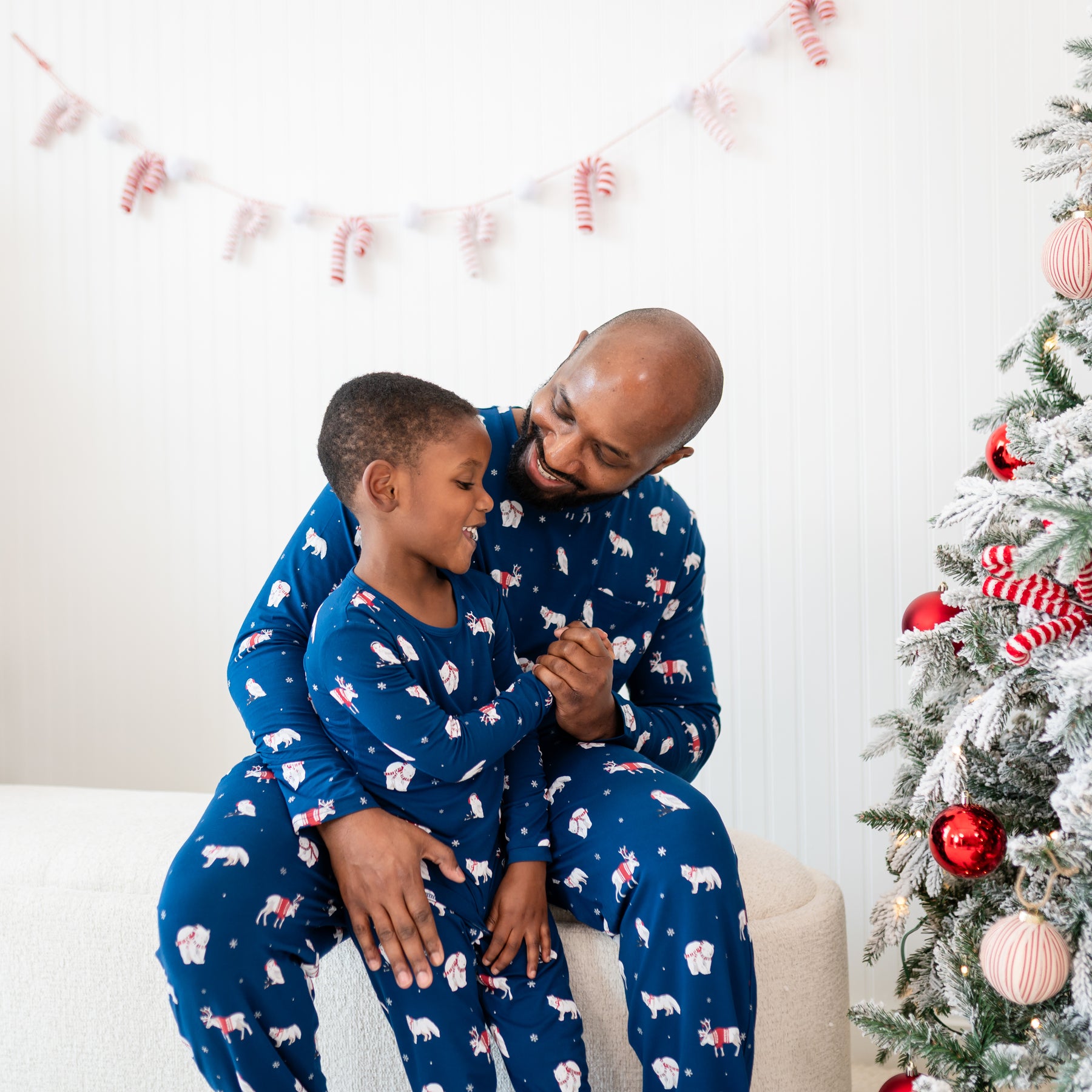 Father sitting on a cream ottoman wearing the Men's Jogger Set in Frosty Friends with his son in front of him who is wearing matching long sleeve toddler pajamas