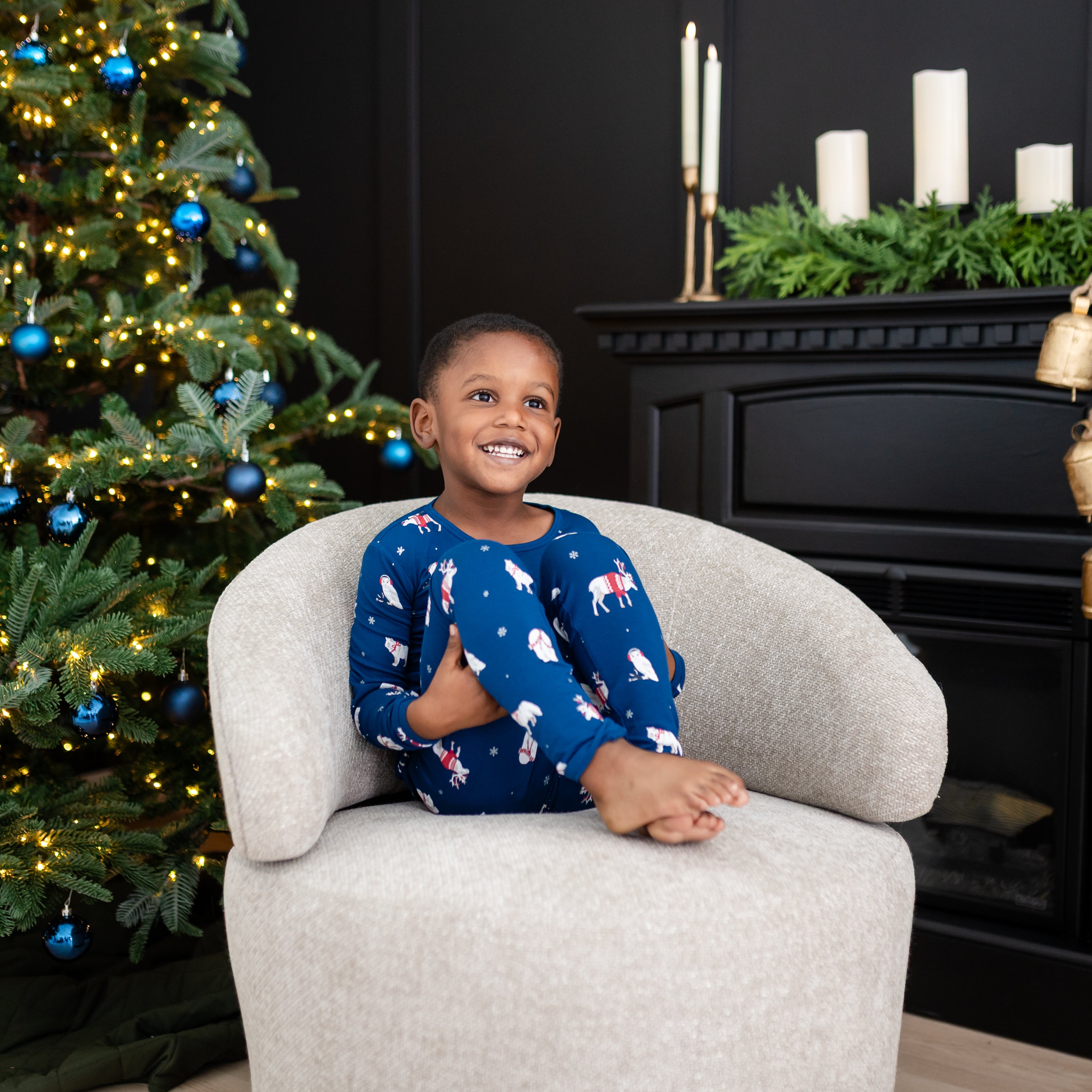 Young smiling boy sitting on a cushioned chair wearing the Long Sleeve Pajamas in Frosty Friends with a decorated Christmas tree and black fireplace behind him