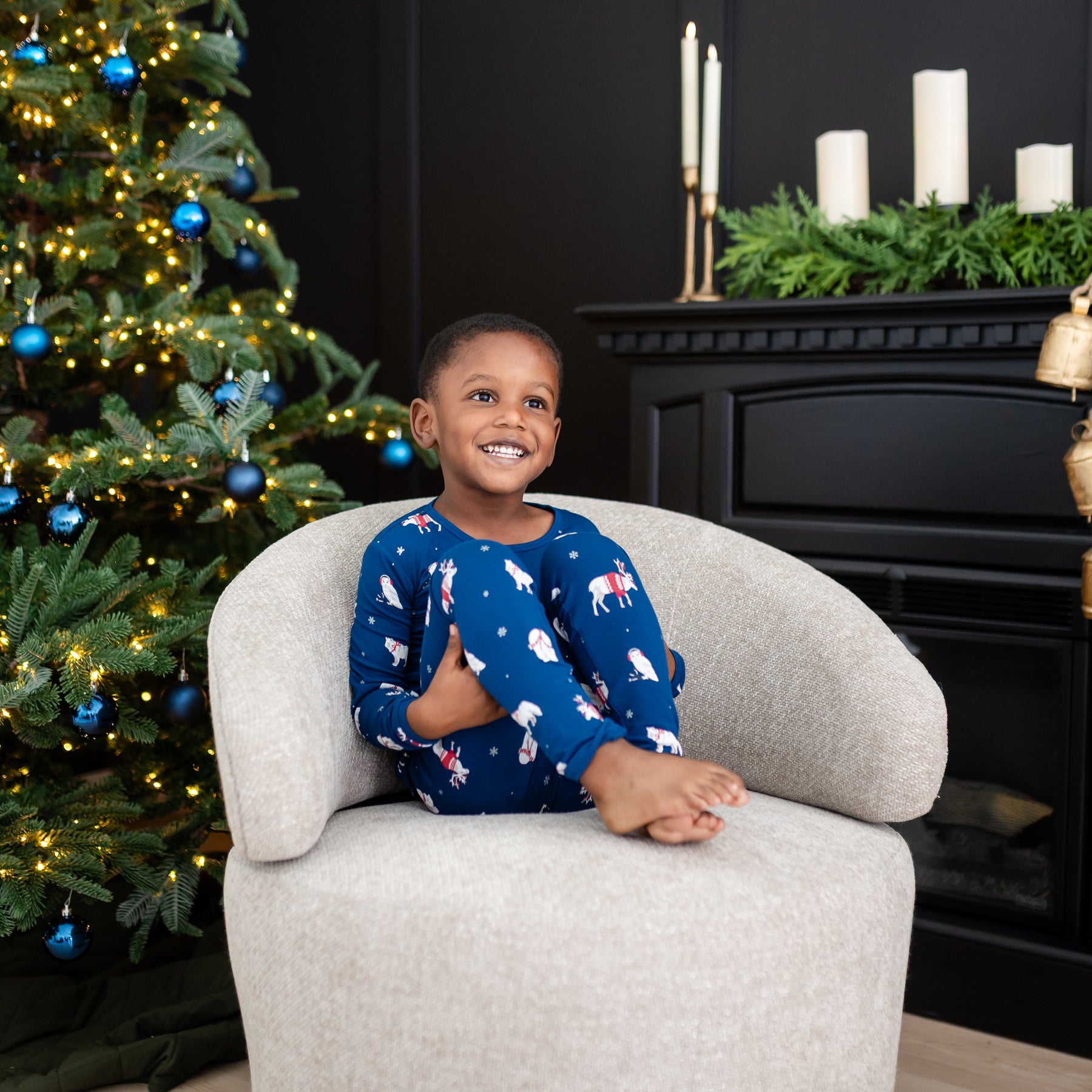 Young smiling boy sitting on a cushioned chair wearing the Long Sleeve Pajamas in Frosty Friends with a decorated Christmas tree and black fireplace behind him