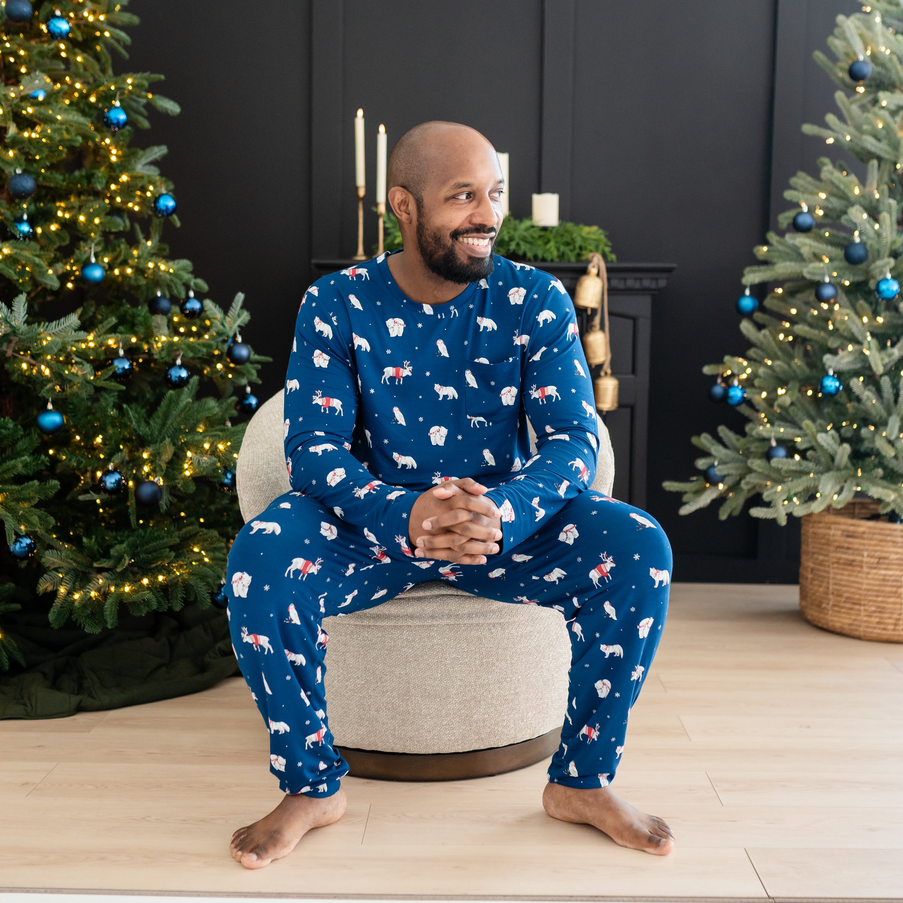 Male model sitting on a cushioned chair wearing the Men's Jogger Set in Frosty Friends in front of two decorated Christmas trees and a black fireplace