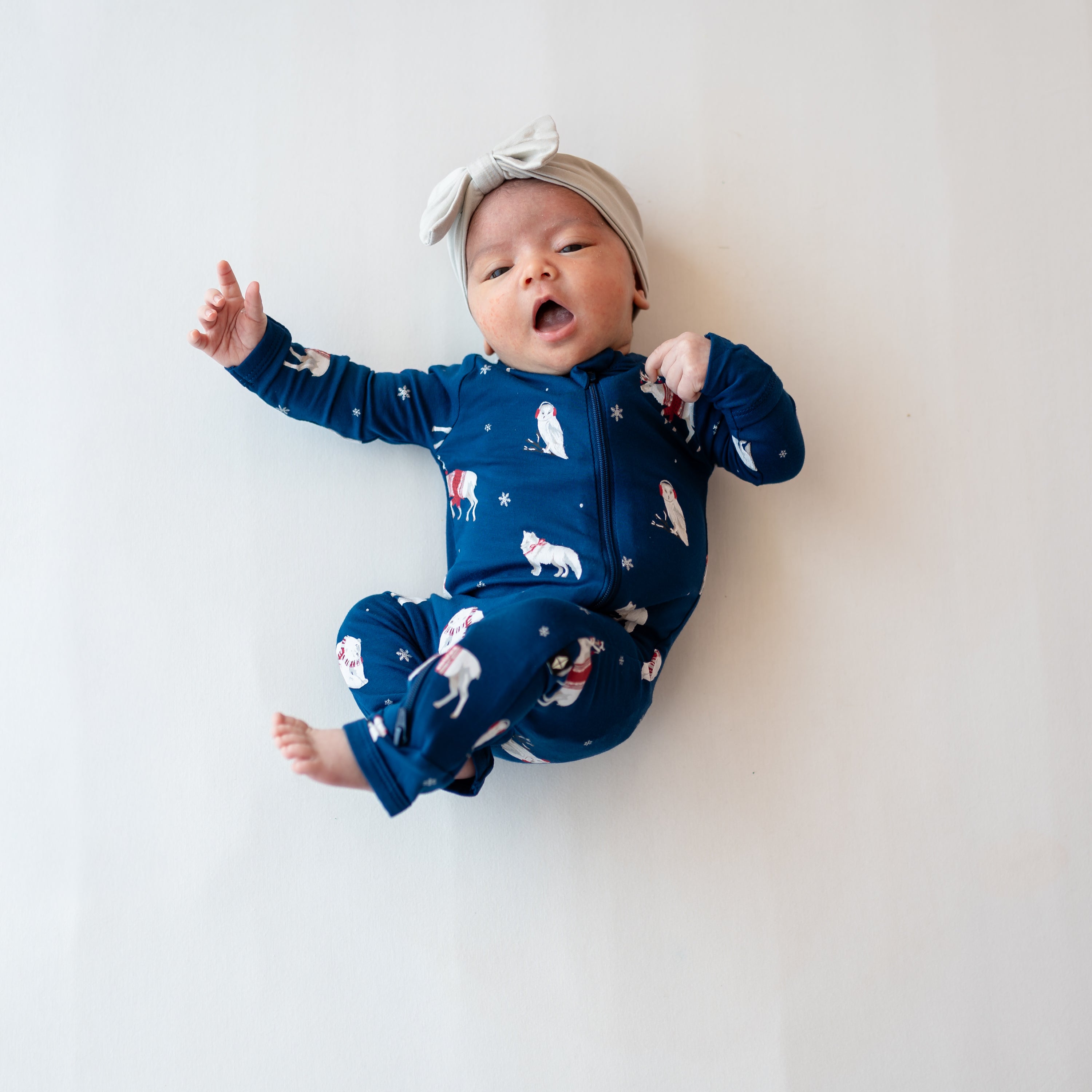 Yawning newborn wearing the Zippered Romper in Frosty Friends laying on a light neutral colored blanket