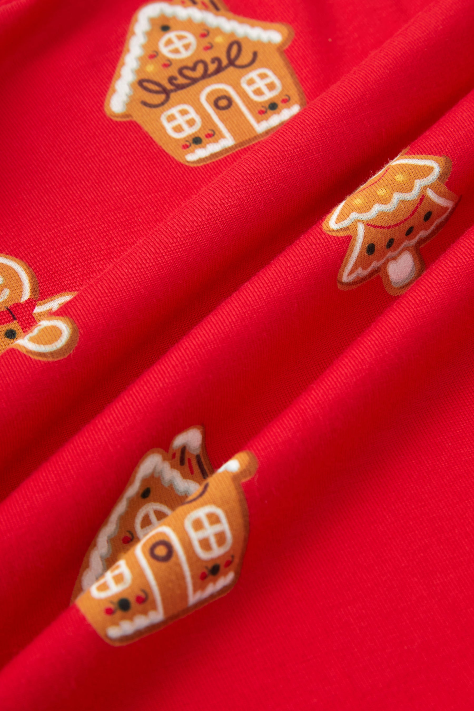 A close-up of bright red fabric with repeating gingerbread house and tree motifs in orange, white, and brown.