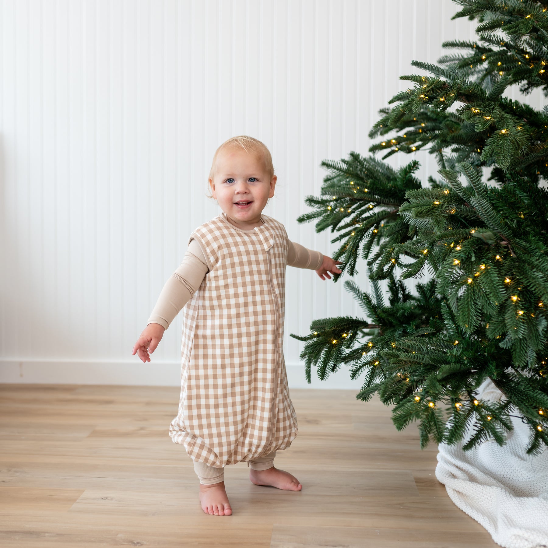 Toddler standing by a Christmas tree wearing the Sleep Bag Walker in Gingham Bisque 1.0 with Bisque romper on underneath