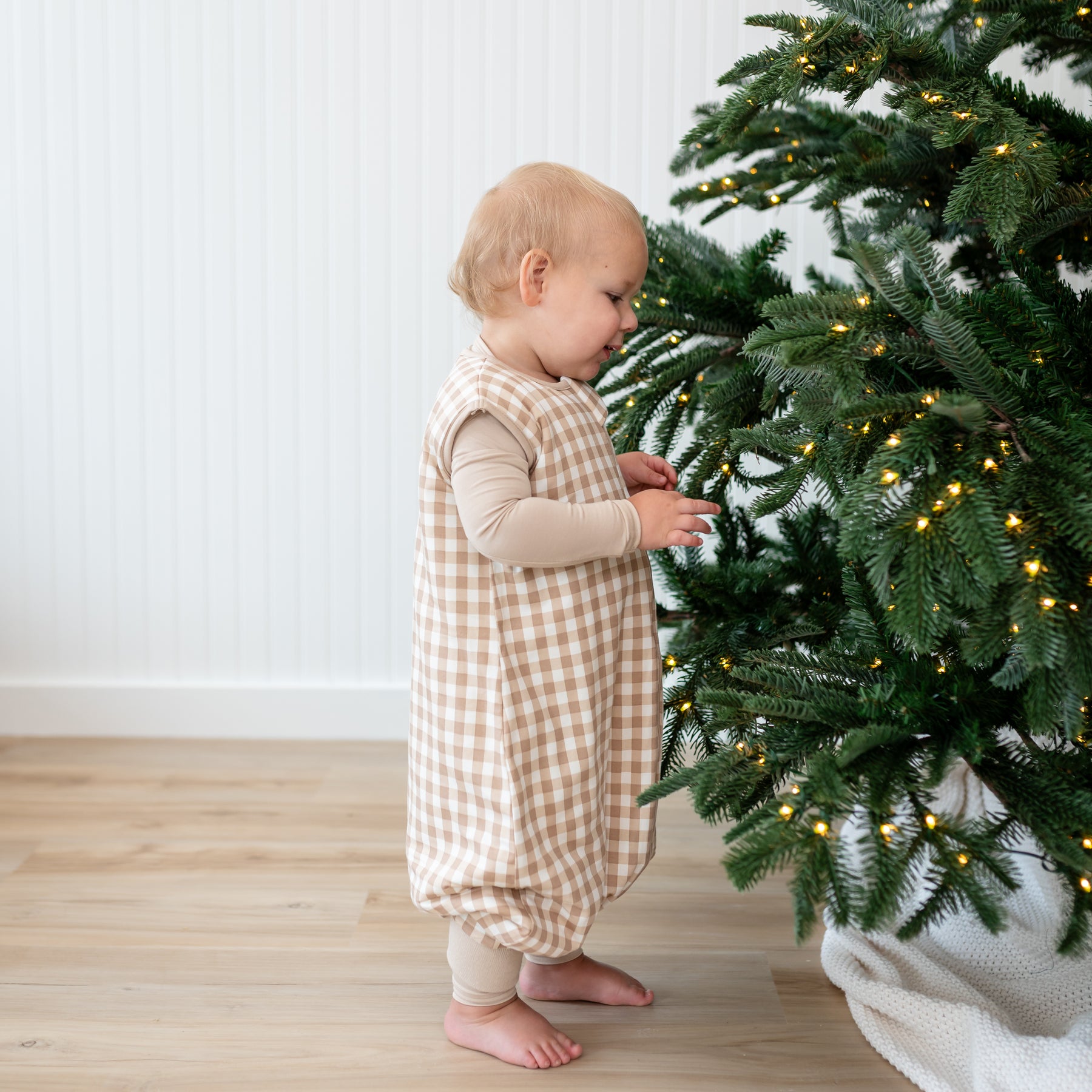 Toddler standing beside a Christmas Tree wearing the Sleep Bag Walker in Gingham Bisque 1.0