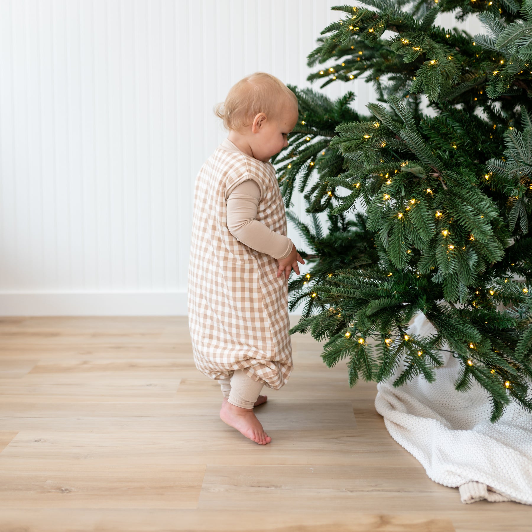 Toddler walking wearing the Sleep Bag Walker in Gingham Bisque 1.0 beside a Christmas Tree