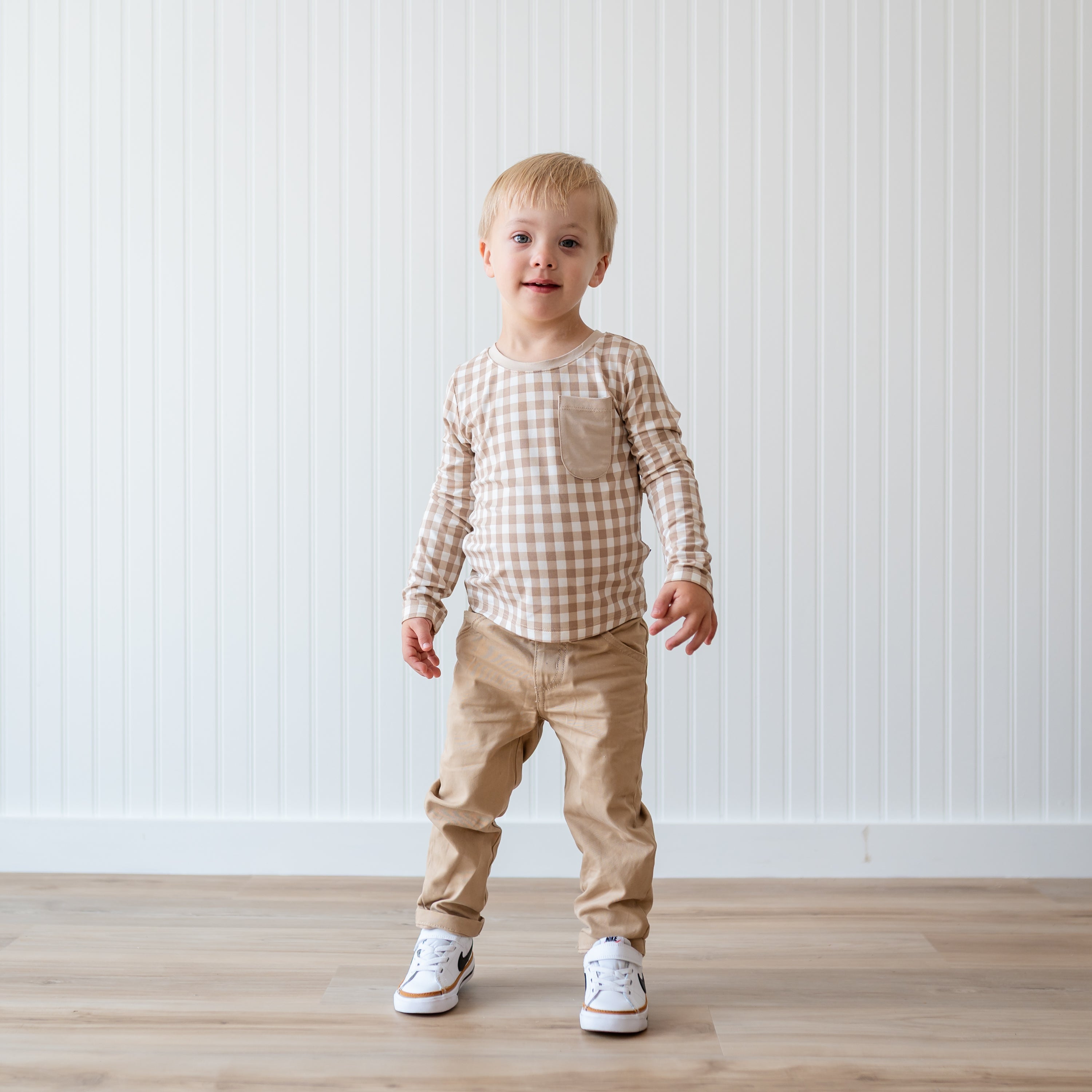 Young boy wearing the Long Sleeve Toddler Crew Neck Tee in Gingham Bisque paired with khaki colored pants standing in front of a white paneled wall