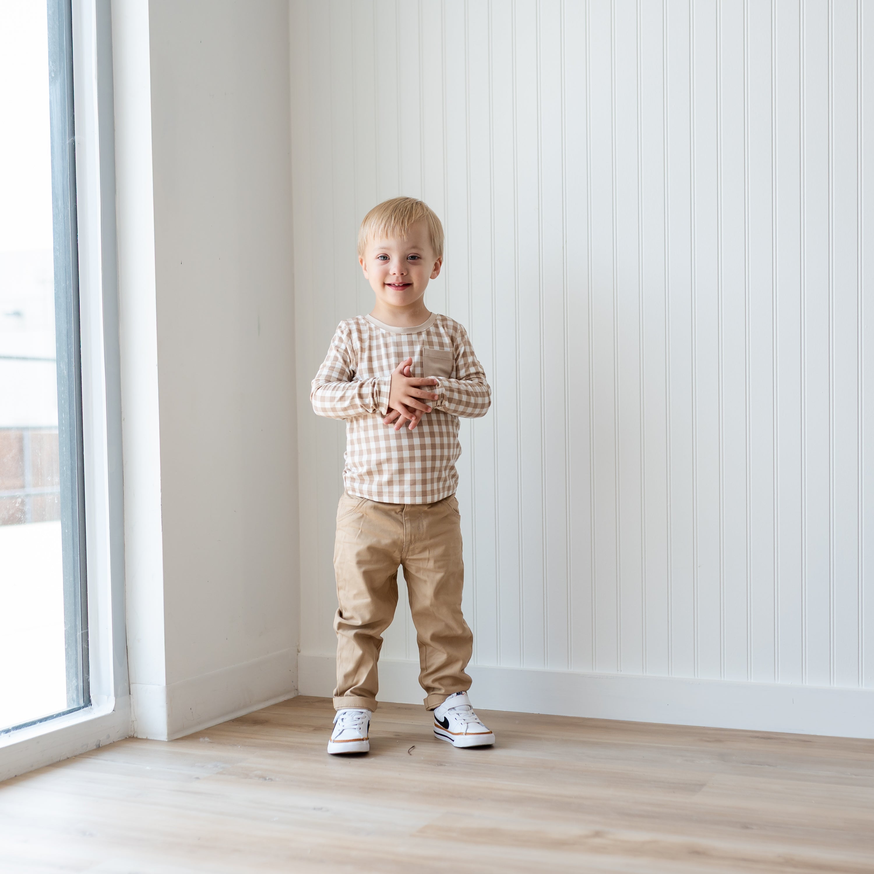 Young boy standing with his hands in front of his chest wearing the Long Sleeve Toddler Crew Neck Tee in Gingham Bisque paired with Khaki colored pants in front of a white paneled wall