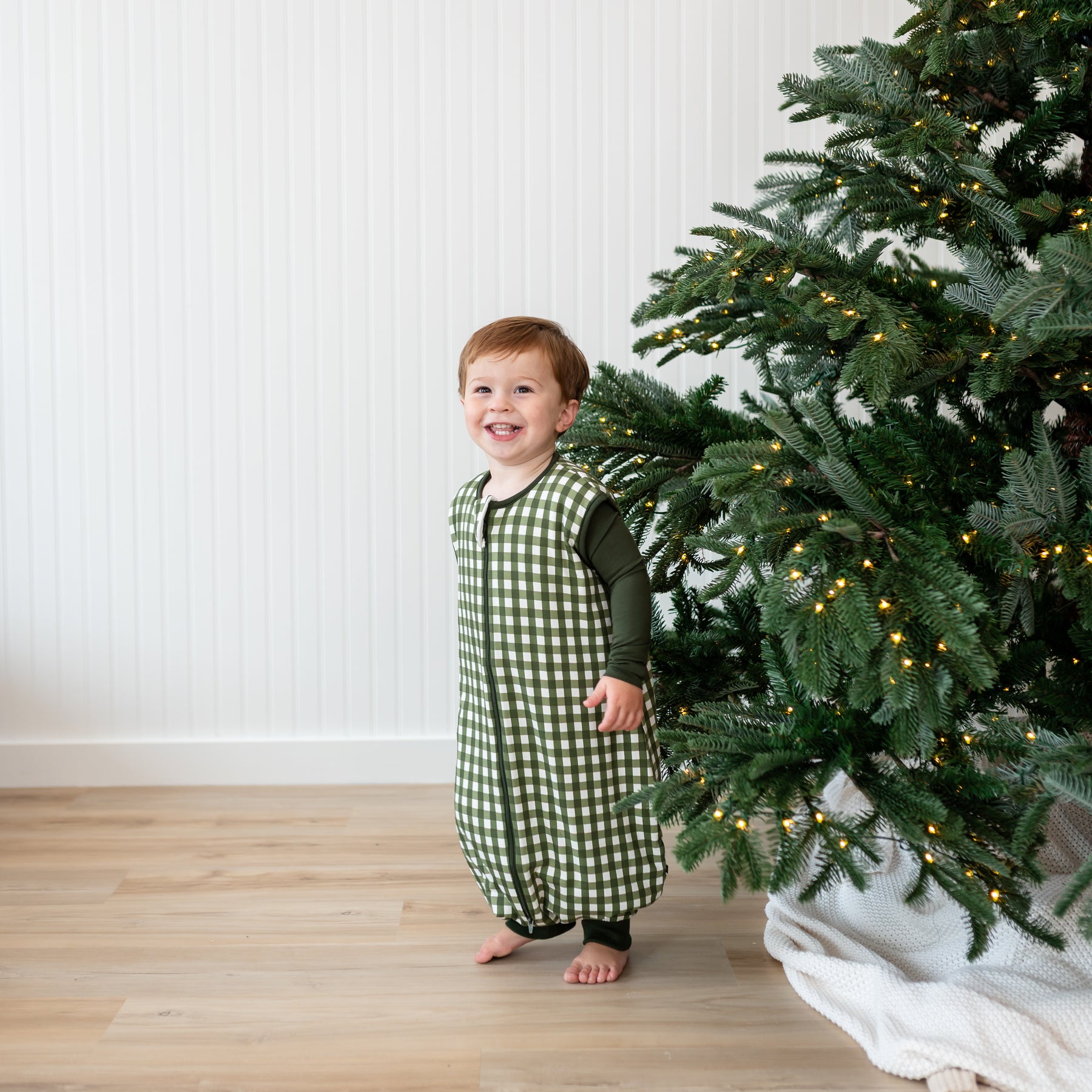 Smiling toddler standing beside a Christmas tree wearing the Sleep Bag Walker in Gingham Fir 1.0