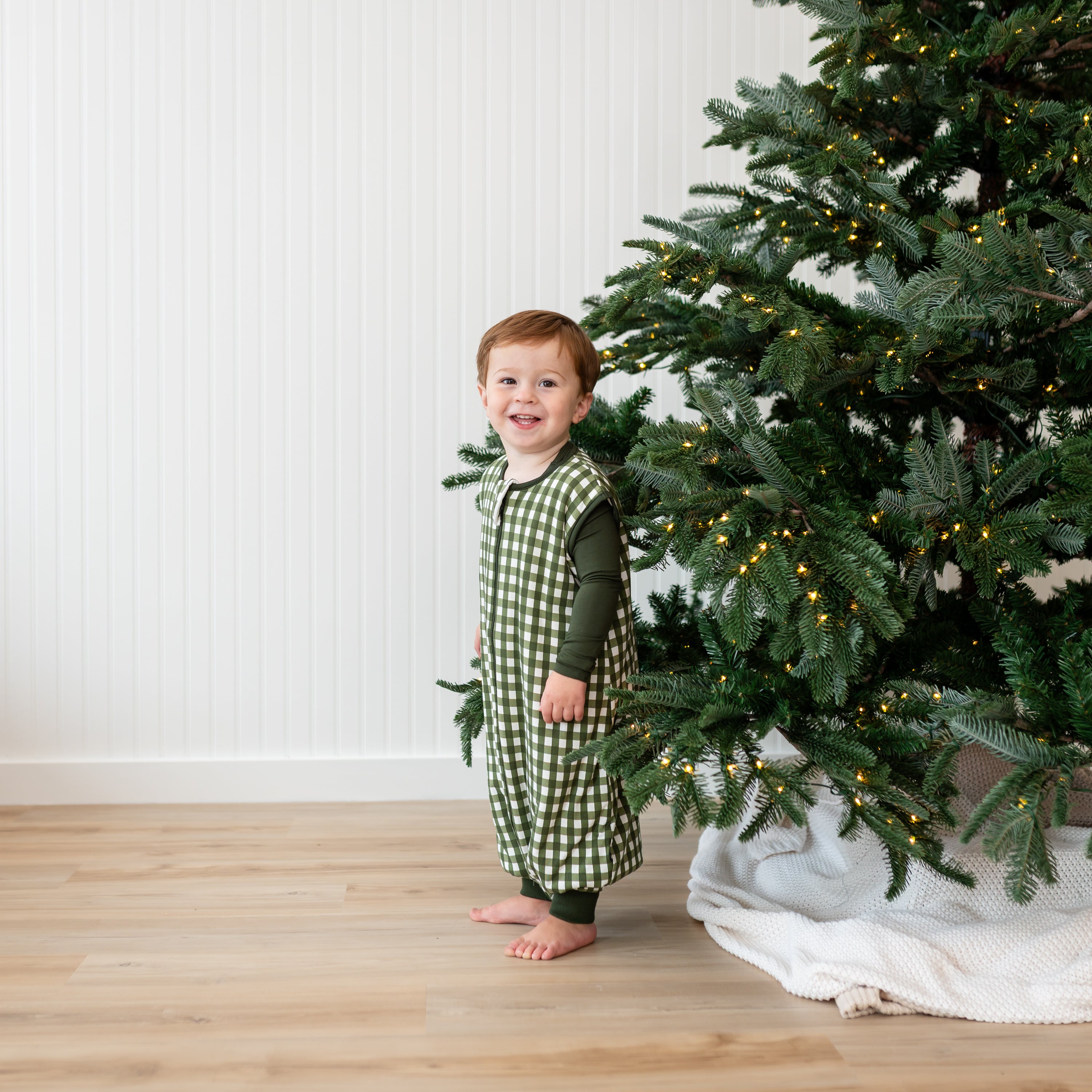 Smiling toddler standing beside a Christmas Tree wearing the Sleep Bag Walker in Gingham Fir 1.0