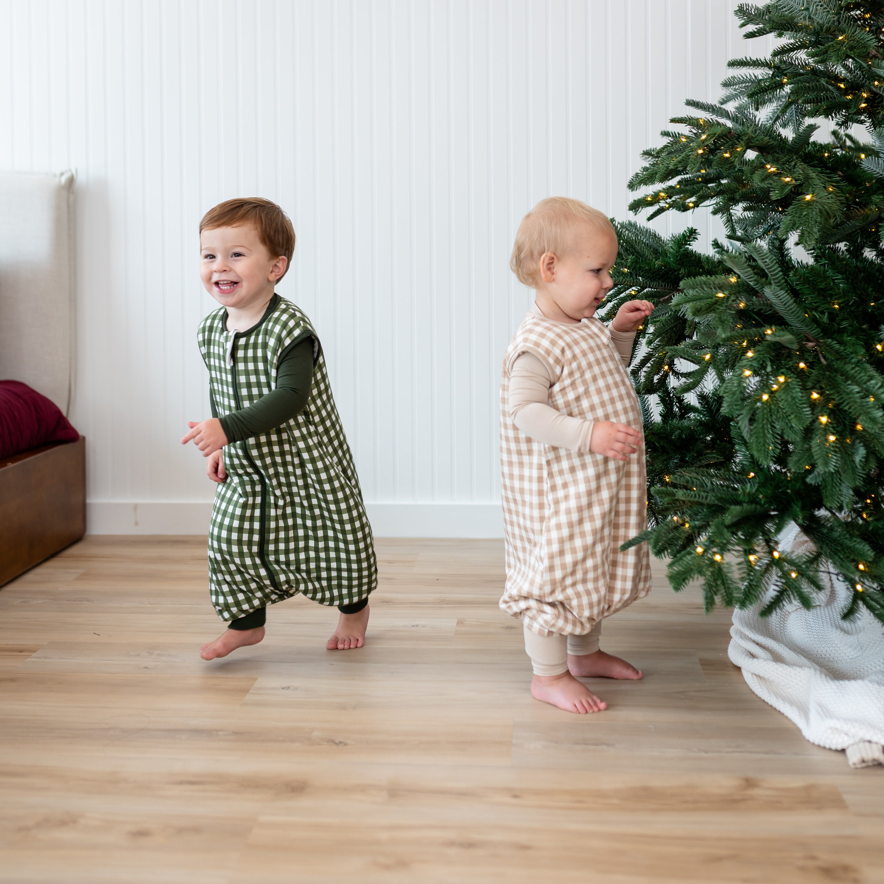 Two toddlers standing beside a Christmas tree wearing the Sleep Bag Walker in Gingham Fir 1.0 and Gingham Bisque