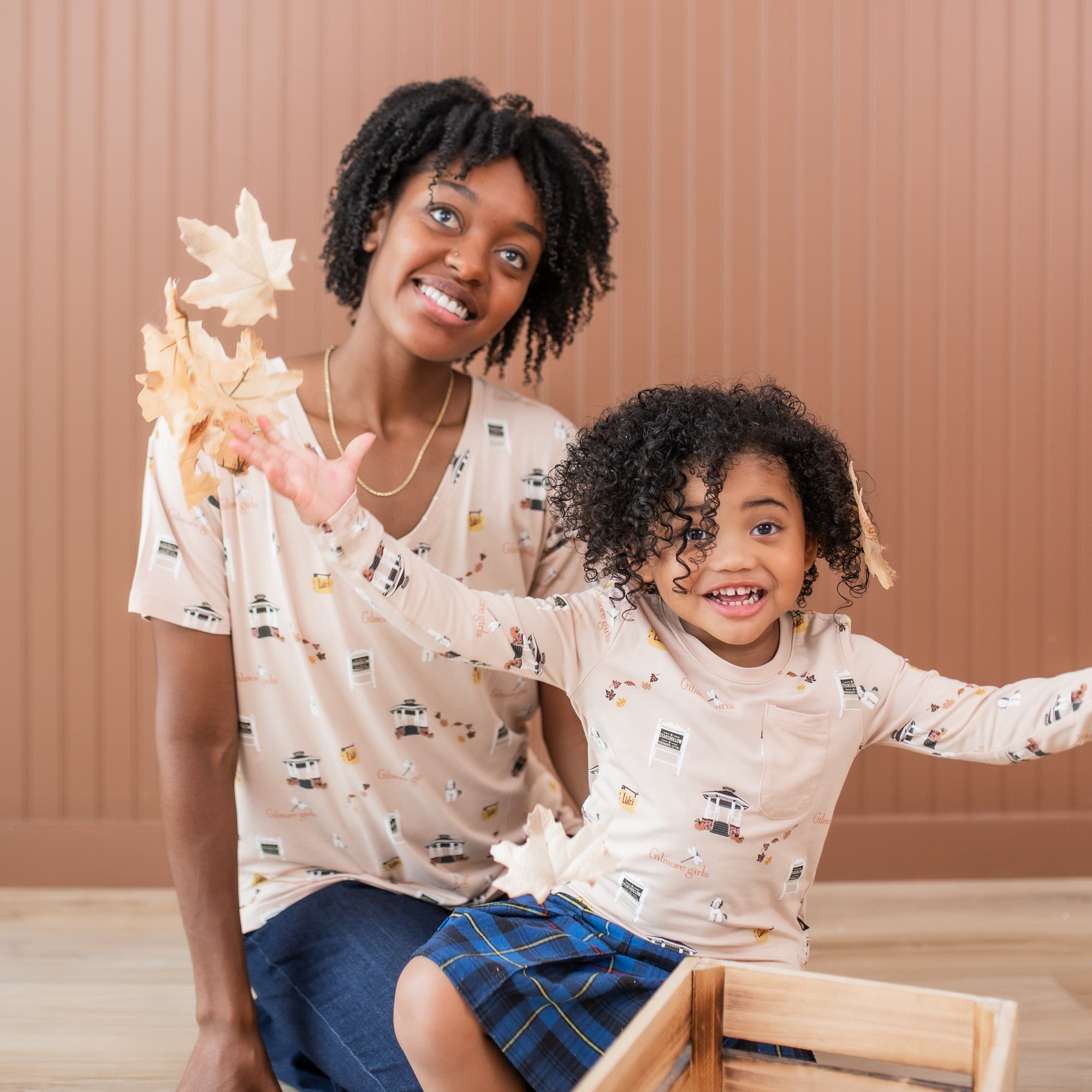Mother and daughter sitting on the floor wearing matching Gilmore Girls themed shirts while daughter throws leaves in the air