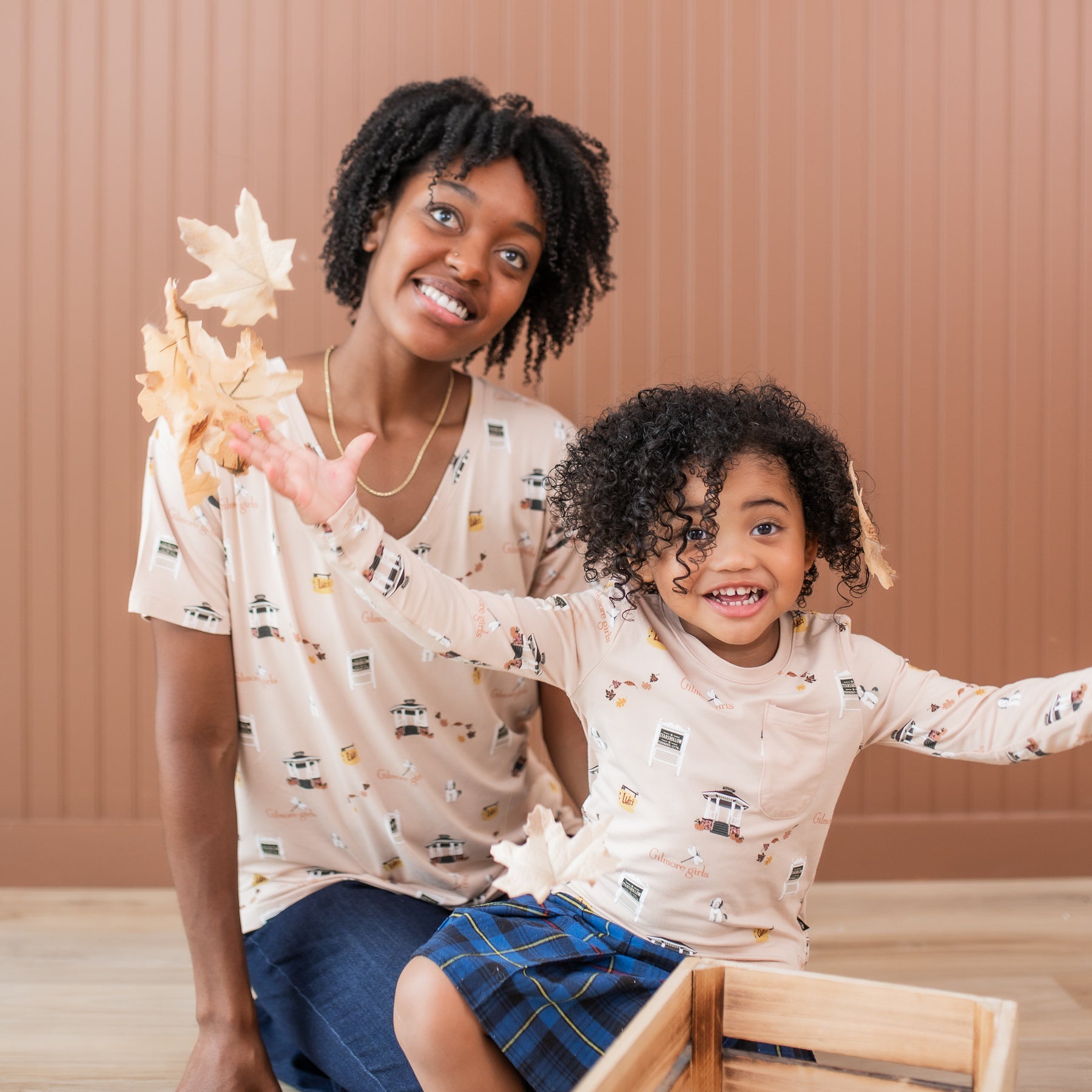 Mother and daughter sitting on the floor wearing matching Gilmore Girls themed shirts while daughter throws leaves in the air