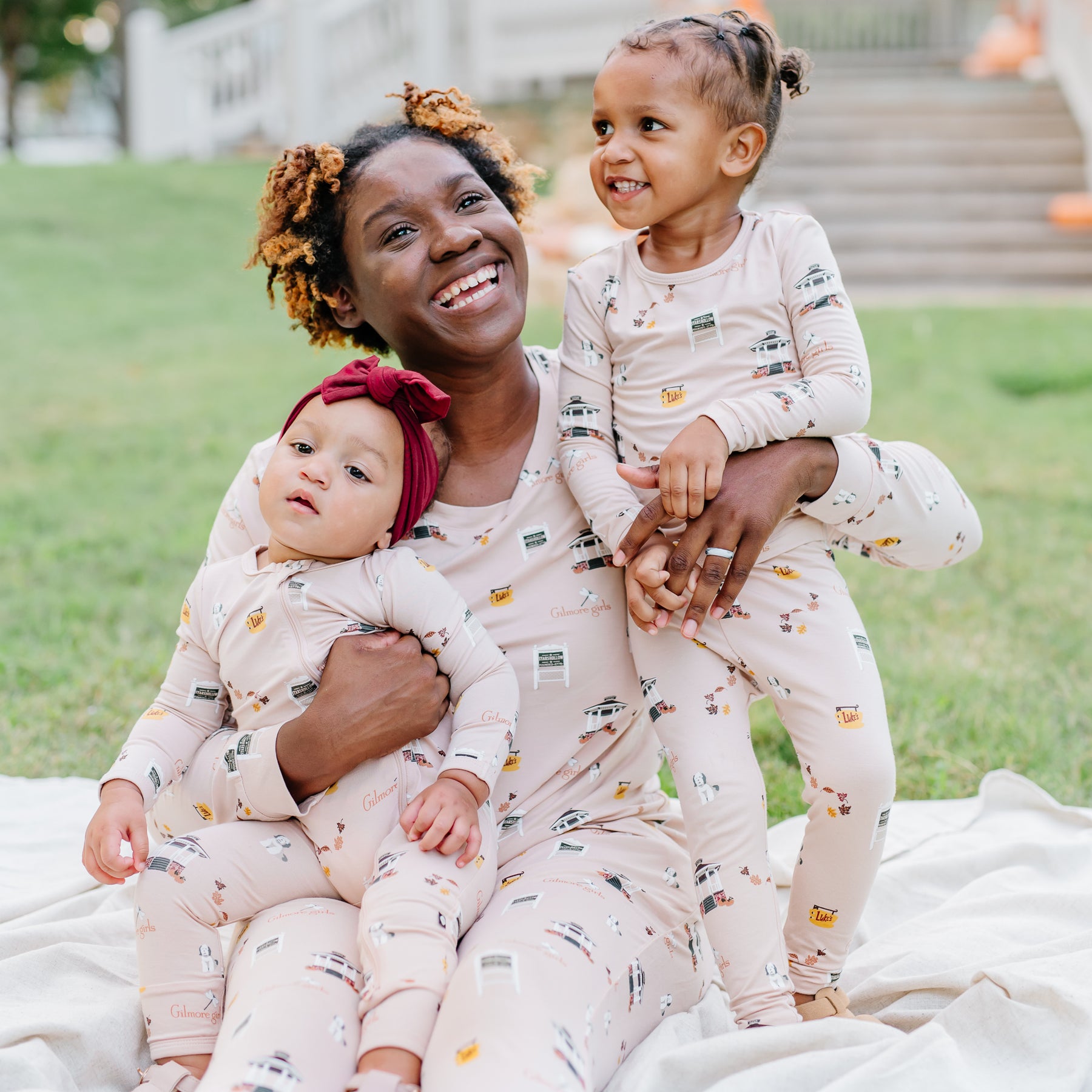 Mother and daughter sitting in the grass on a cream colored blanket wearing various items from the Gilmore Girls collections