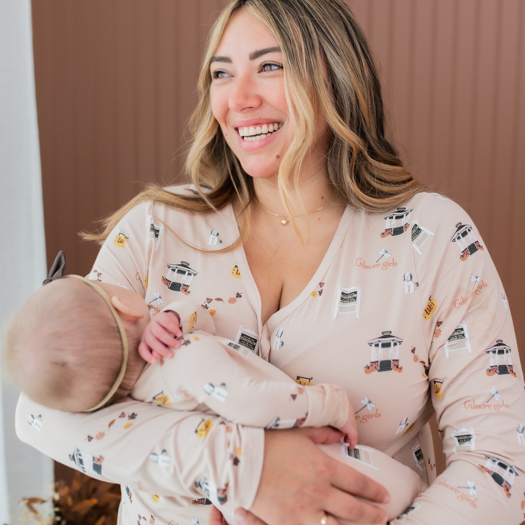 Smiling female model wearing the Women's Long Sleeve Henley Tee in Gilmore Girls holding her infant model wearing the matching zippered romper