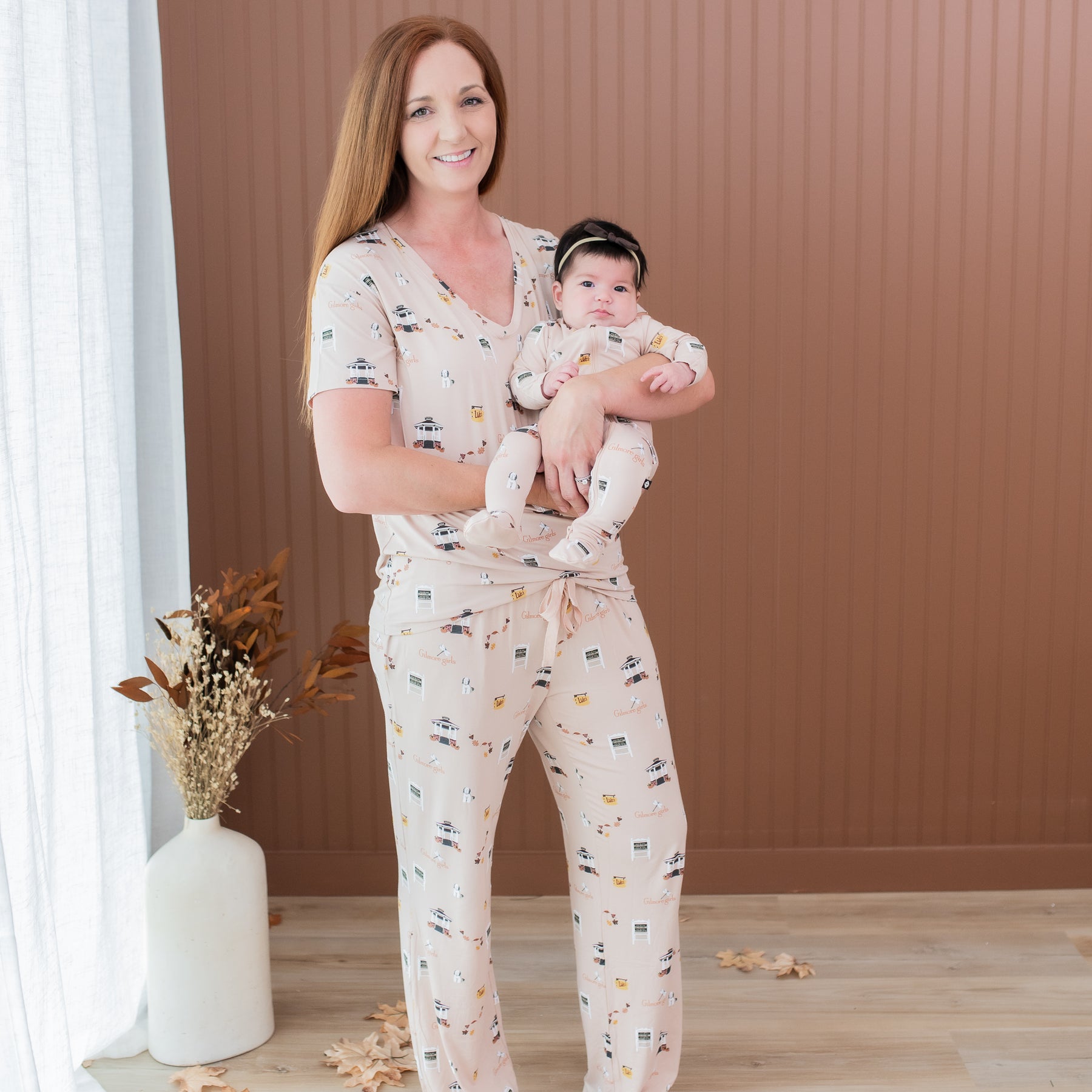 Smiling female model wearing the Women's Lounge Pants in Gilmore Girls with matching Women's V-Neck holding her infant daughter who is wearing a matching zippered footie
