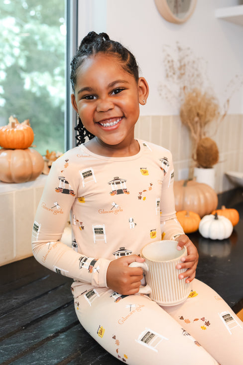 Child in pajamas with Gilmore Girls inspired pattern holding a mug, surrounded by pumpkins indoors.