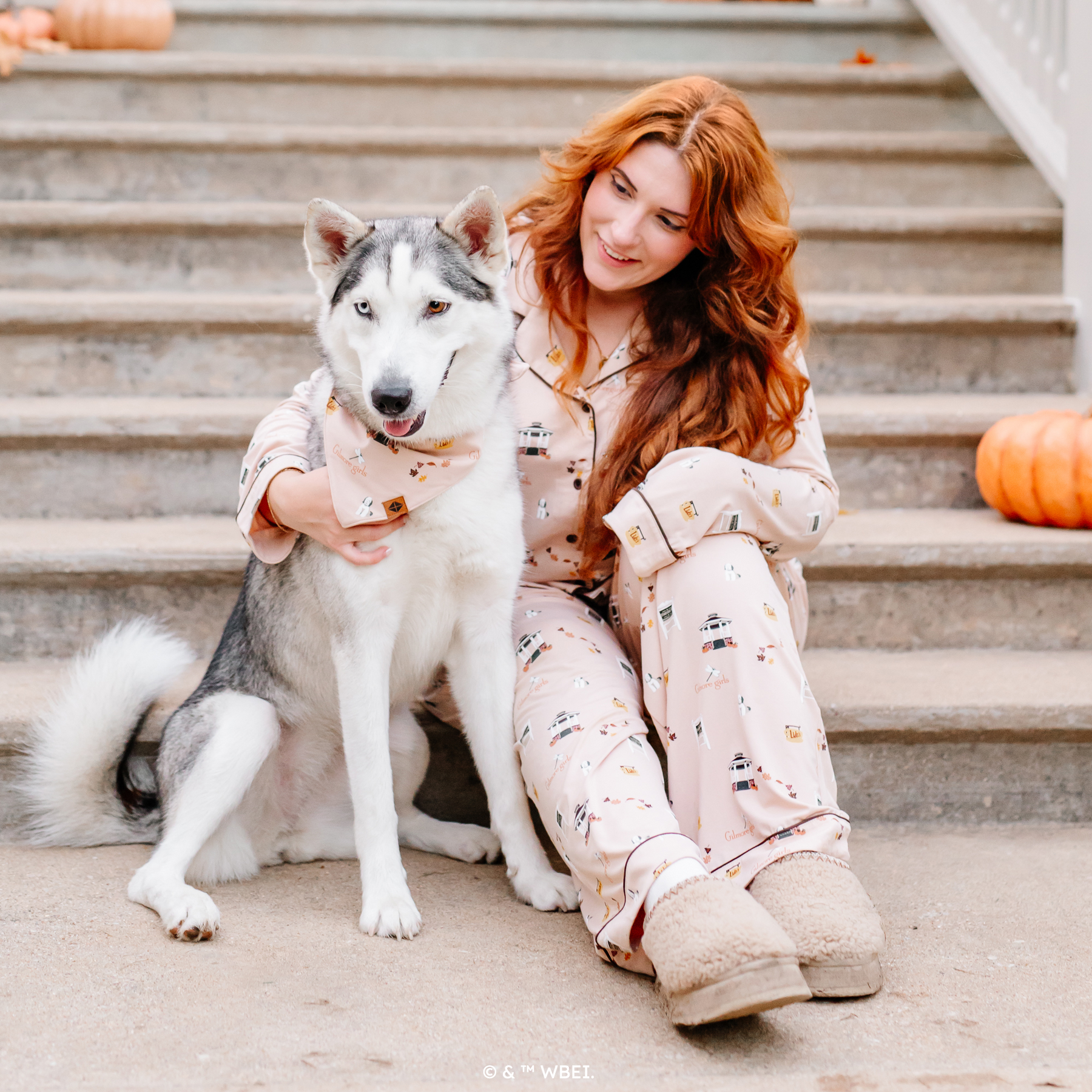 Female model sitting at the bottom of concrete steps of a gazebo wearing the Long-Sleeved Women's Pajama Set in Gilmore Girls with her arm around a husky wearing a matching dog bandana