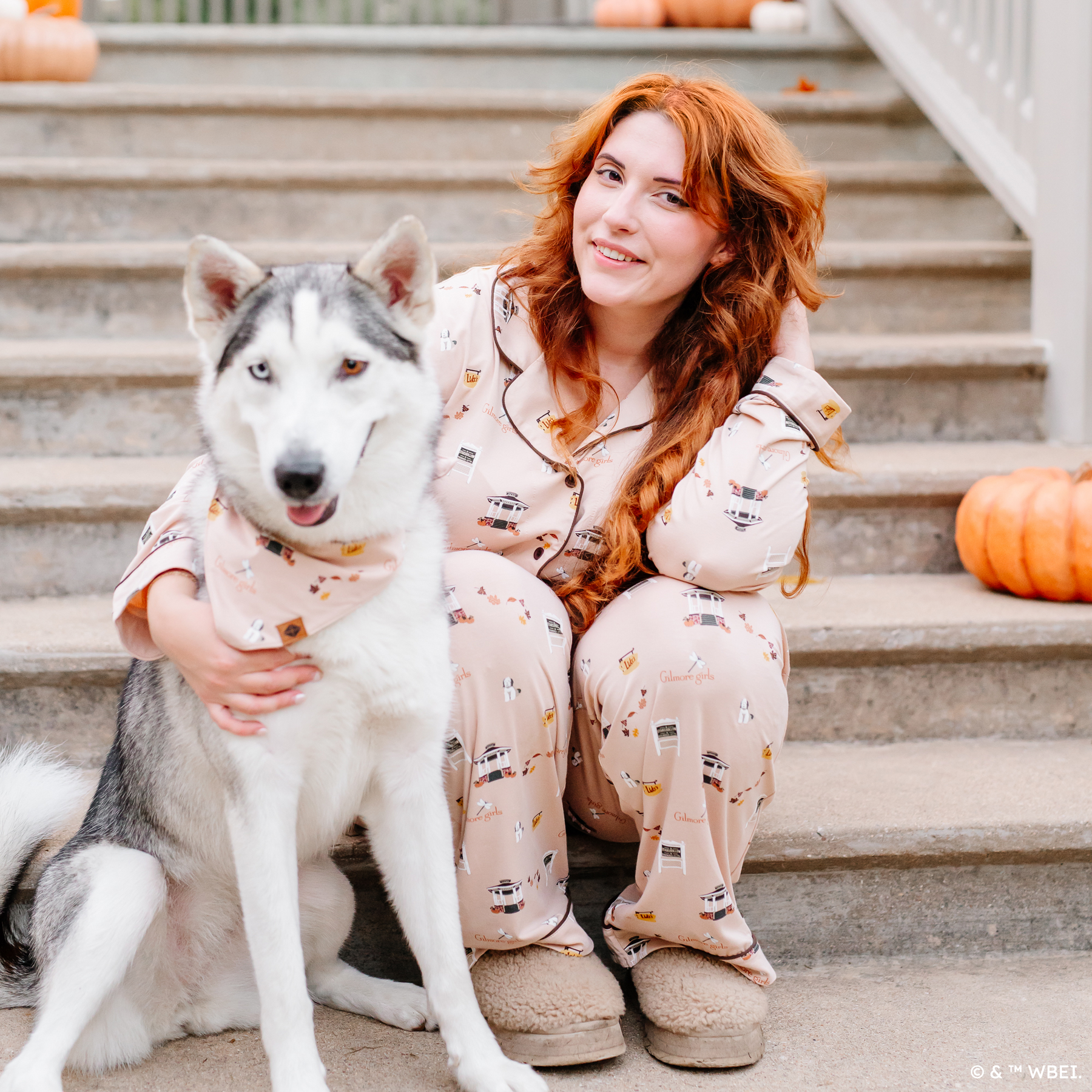 Female model sitting on concrete steps wearing the Long-Sleeved Women's Pajama Set in Gilmore Girls with her arm around a husk dog wearing a matching dog bandana