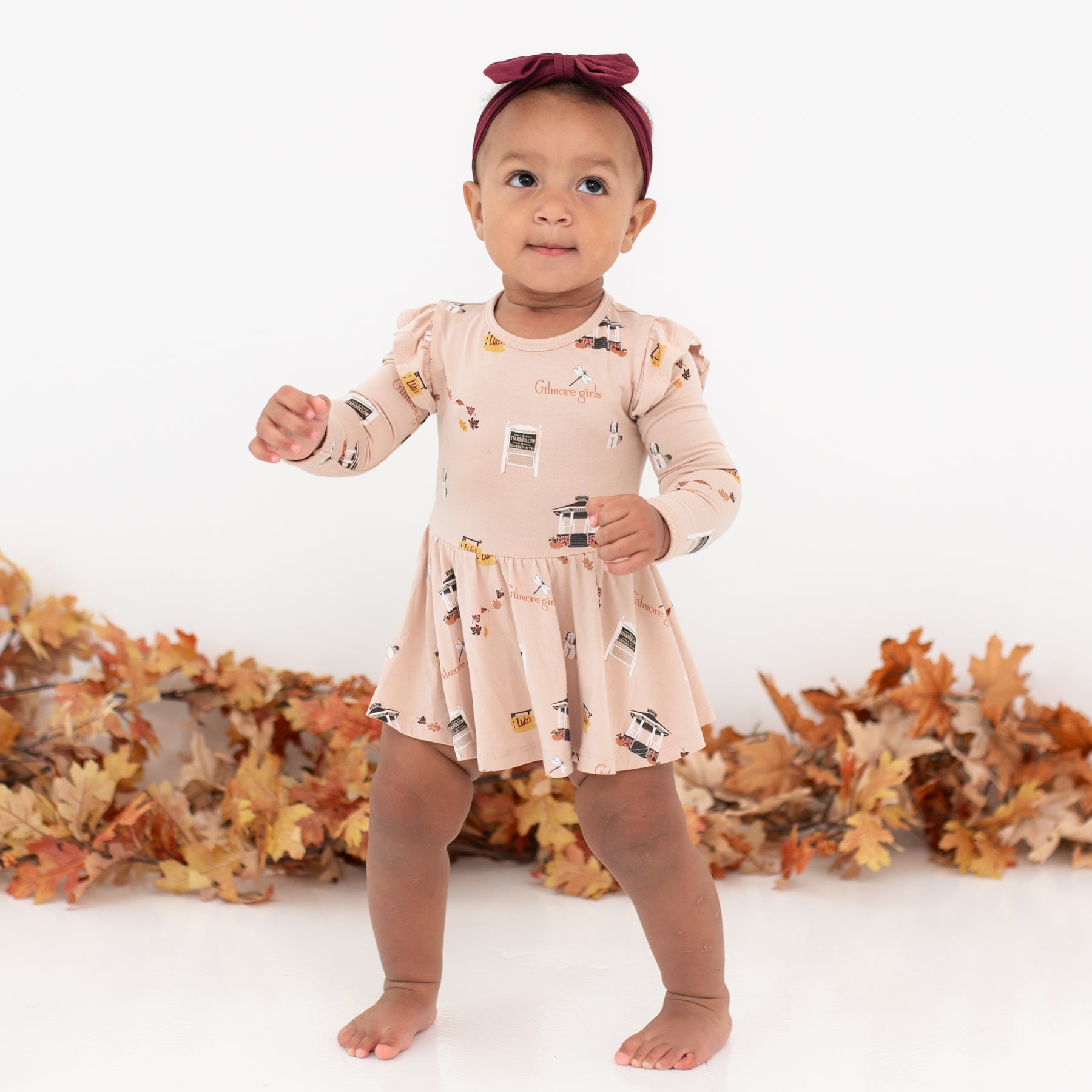 Young toddler walking wearing the Long Sleeve Twirl Bodysuit Dress in Gilmore Girls with a burgundy bow in front of a leaf garland