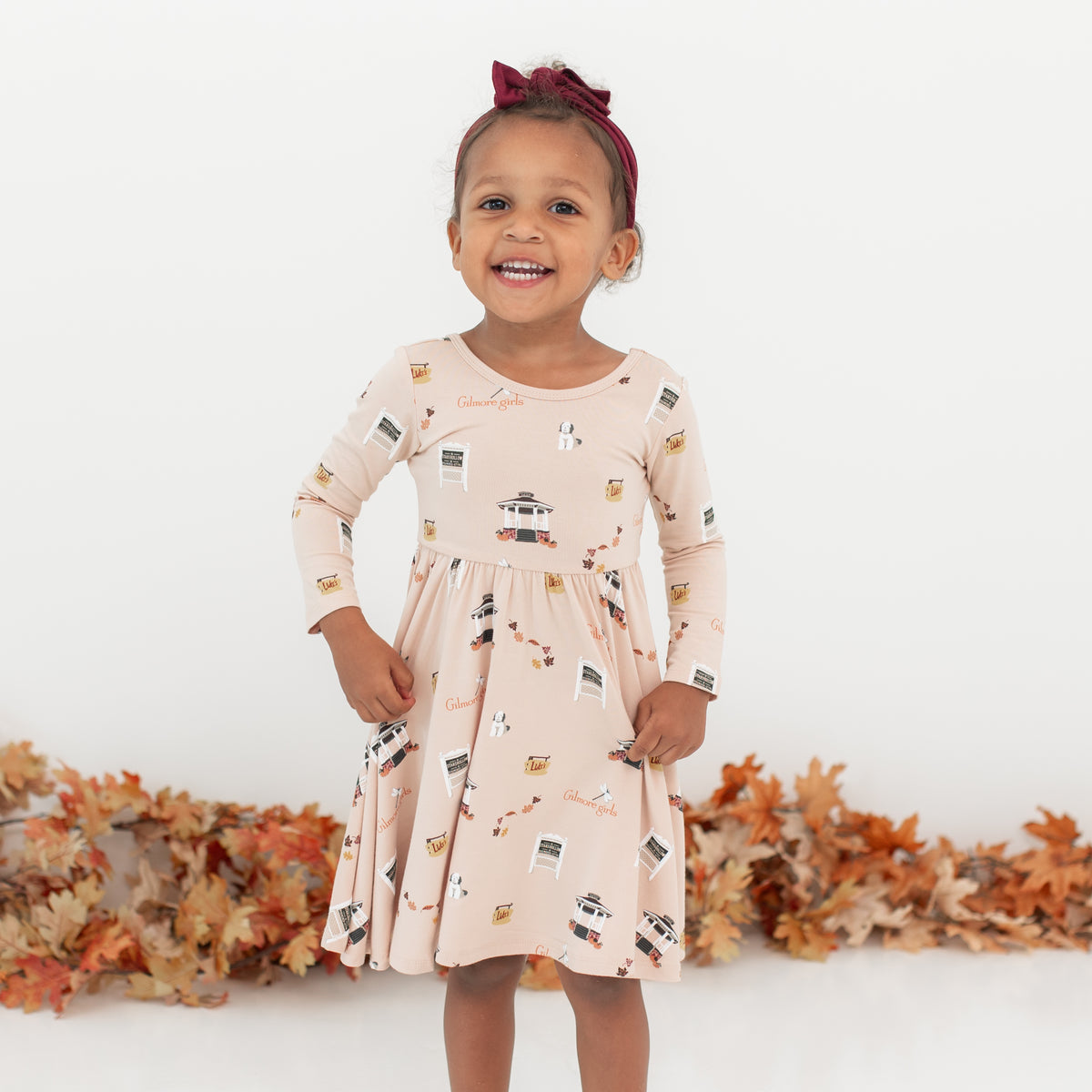 Young girl wearing the Long Sleeve Twirl Dress in Gilmore Girls with a burgundy bow in front of a white backdrop and a leaf garland