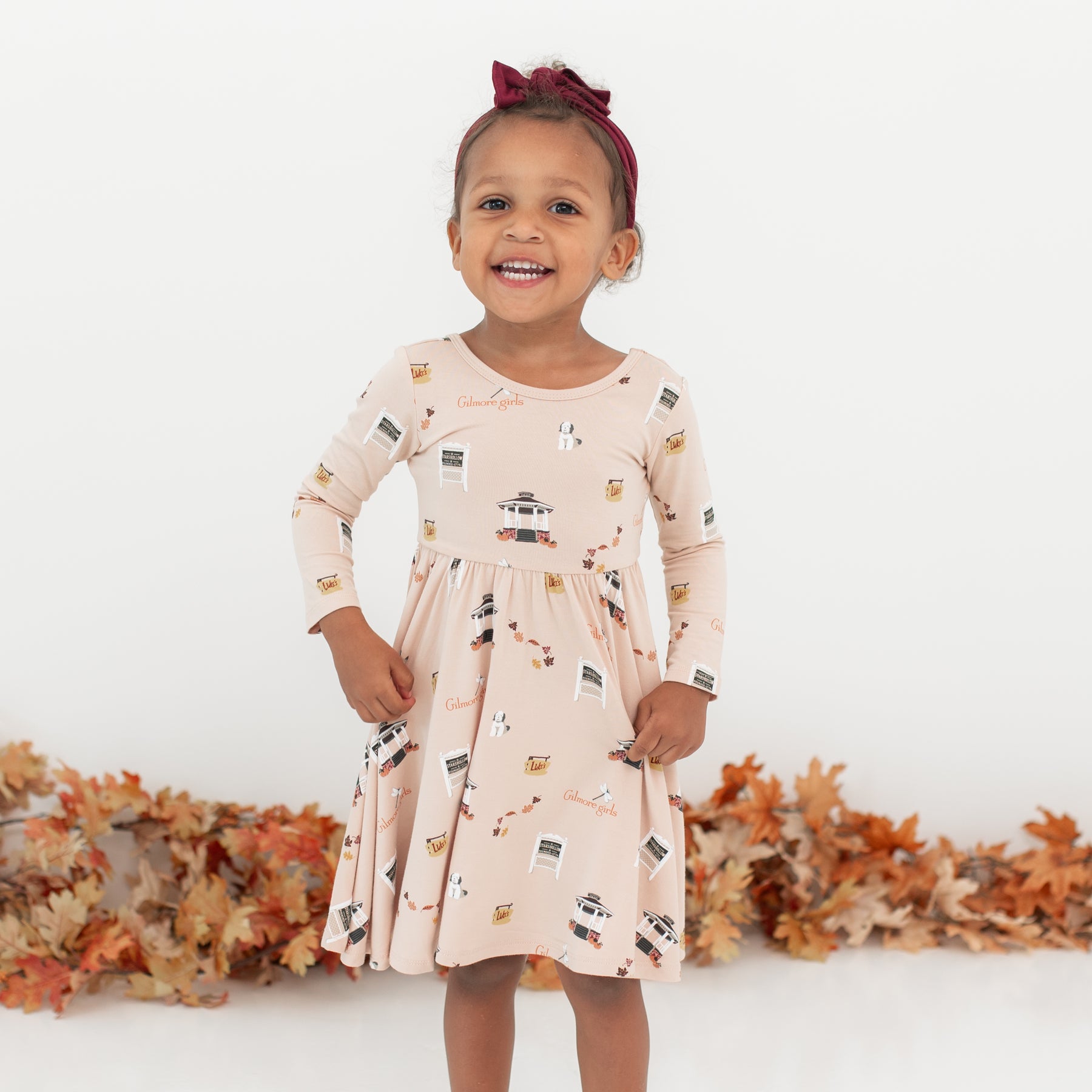 Young girl wearing the Long Sleeve Twirl Dress in Gilmore Girls with a burgundy bow in front of a white backdrop and a leaf garland