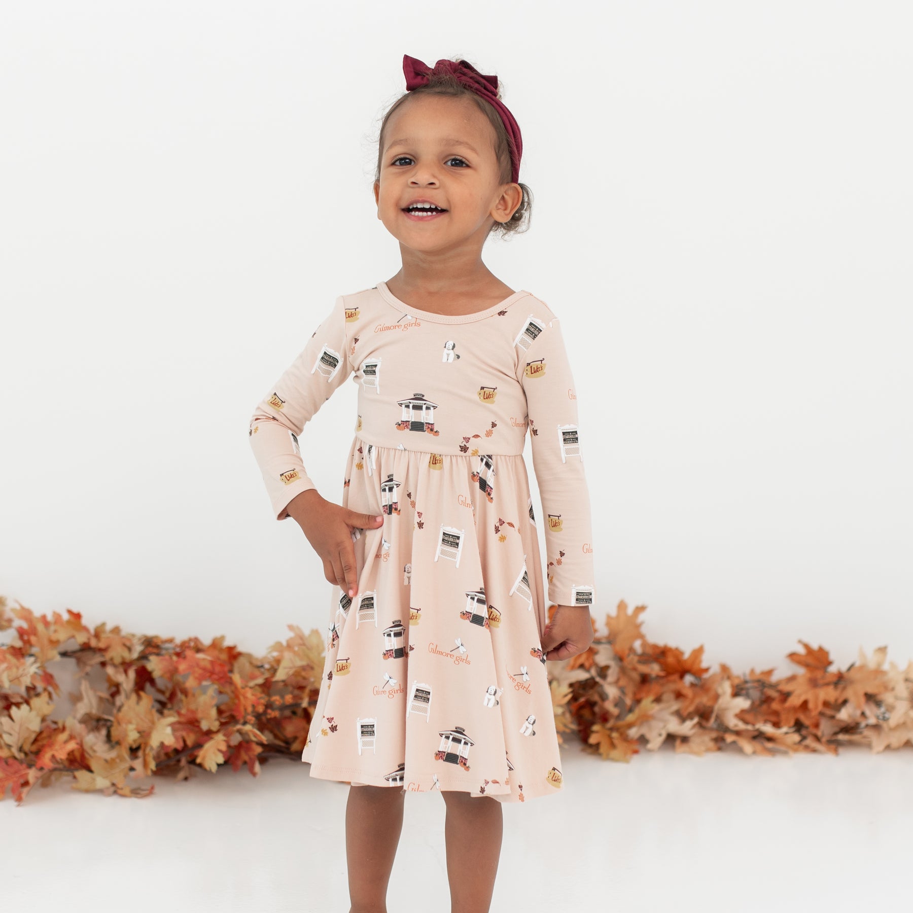 Young girl standing in front of a white wall and leaf garland wearing the Long Sleeve Twirl Dress in Gilmore Girls with a burgundy bow