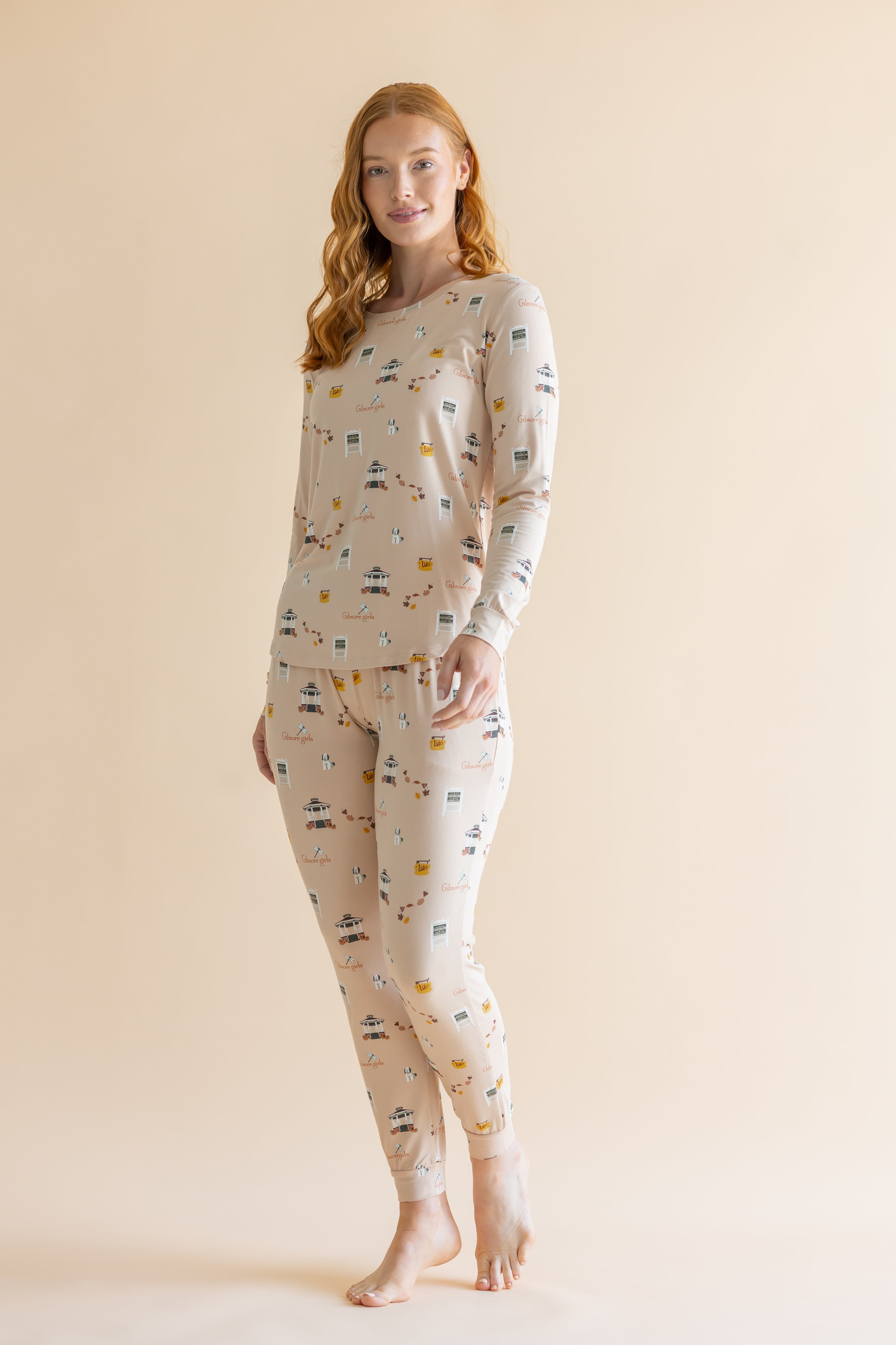 A woman modeling a two-piece long-sleeve pajama set with an all-over Gilmore Girls inspired print on a warm cream background, set against a solid light beige studio backdrop.