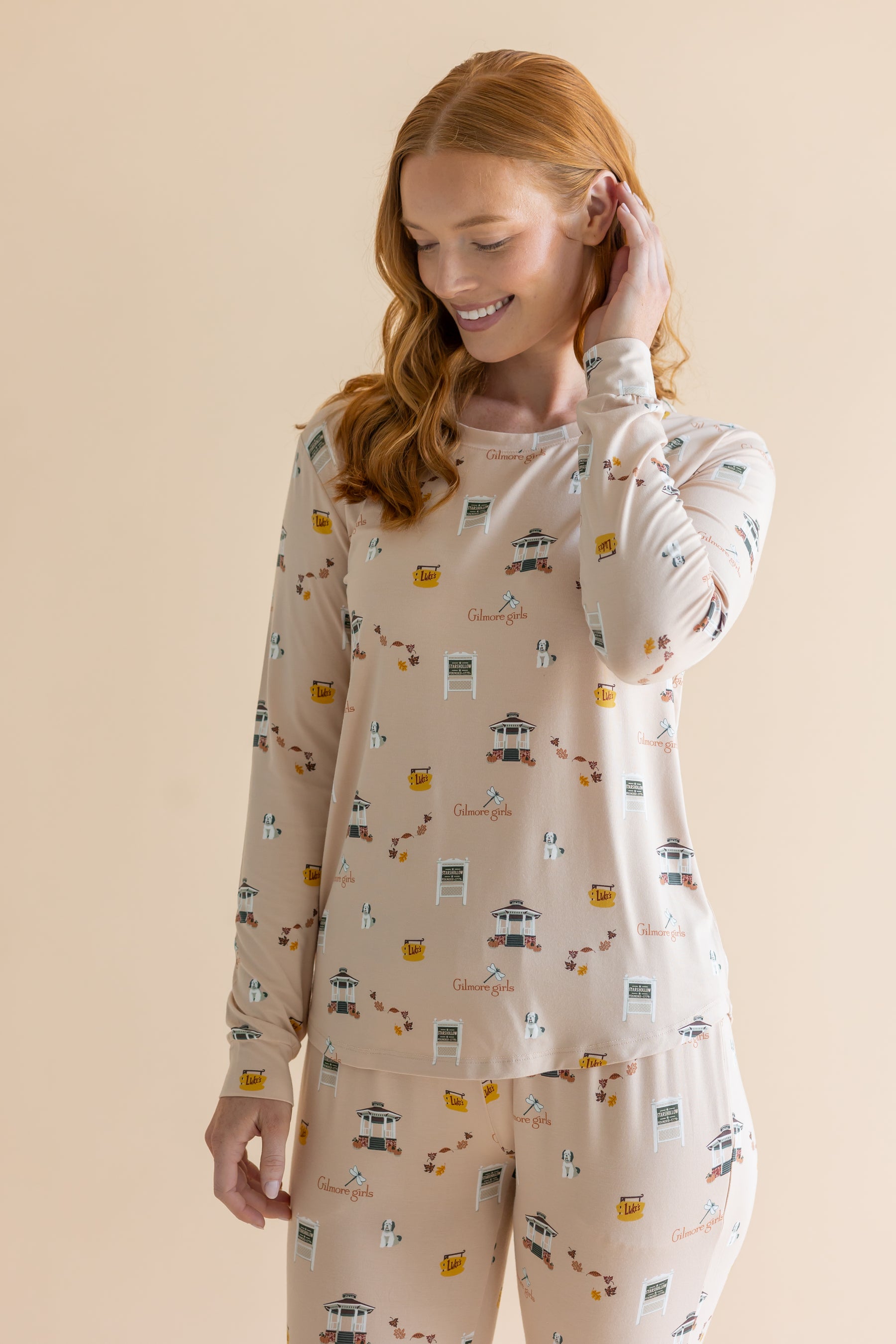 A woman modeling a two-piece pajama set with a long-sleeve top and matching pants in a warm beige color, featuring a small repeating pattern of houses, coffee cups, and the word 'Gilmore Girls'.