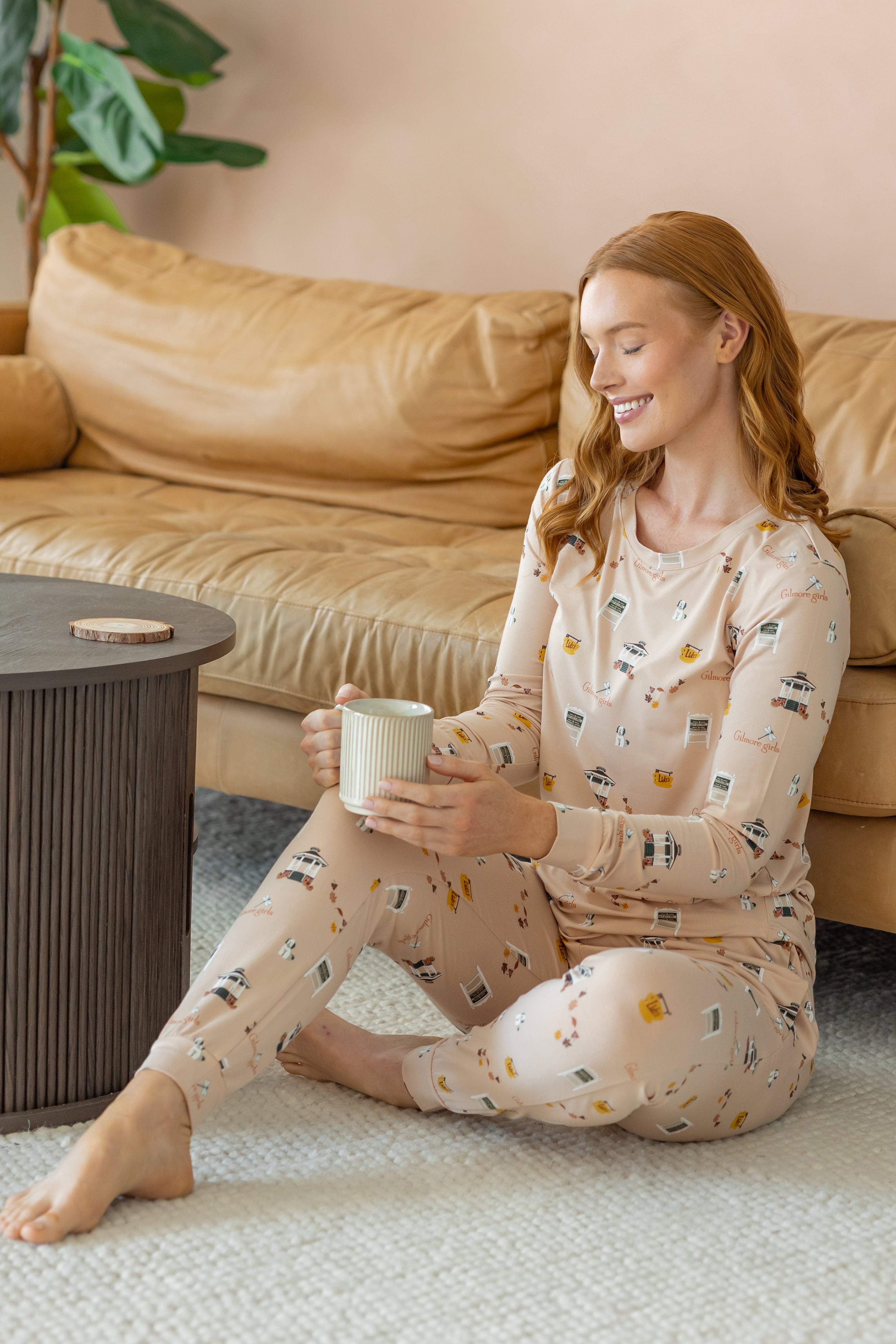 A woman sits on a light rug in a living room setting, wearing a beige long-sleeve pajama set with a small multicolor pattern and holding a white mug.