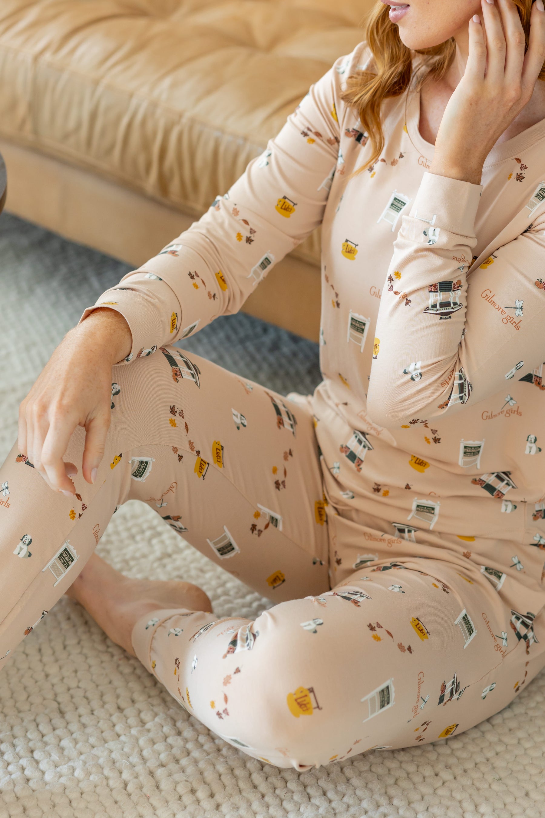 A woman wearing a long-sleeve pajama set with matching pants, sitting on a textured rug in a cozy indoor setting. The pajamas are a light peach-beige color with a small scattered print of  Gilmore Girls inspired elements.