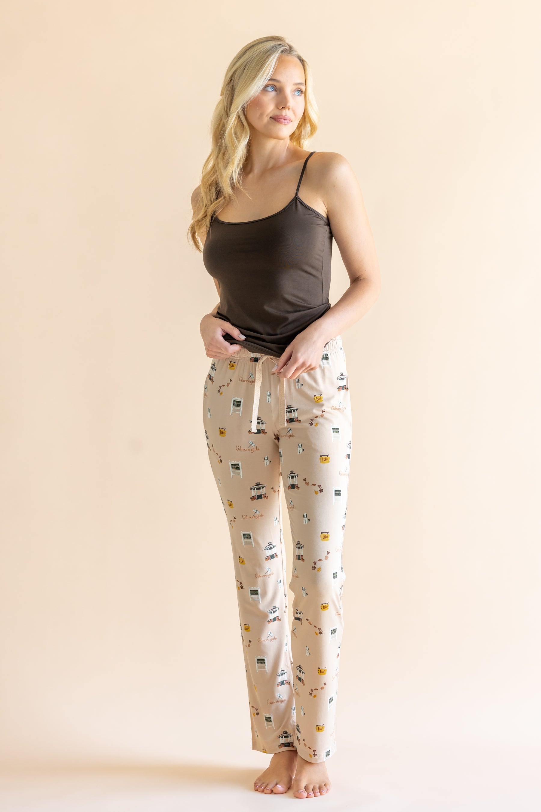 A woman modeling a pajama set consisting of a dark brown camisole top and cream-colored pants with a Gilmore Girls inspired pattern, set against a solid light beige background.