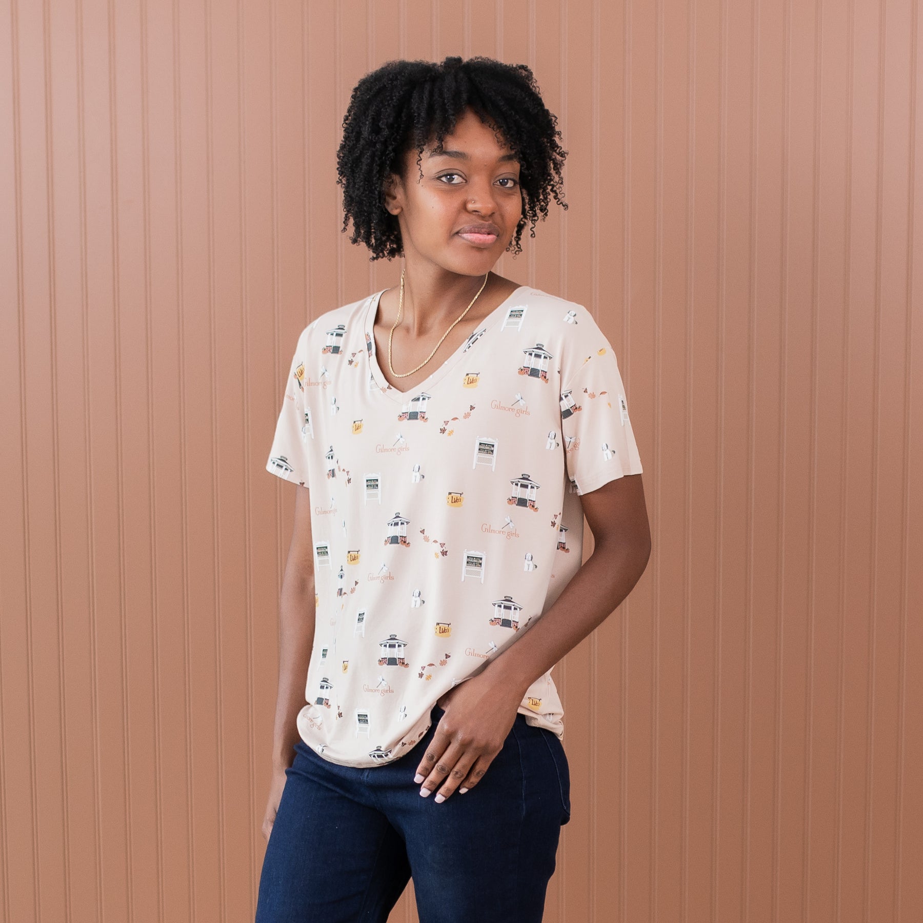 Female modeling the Women's Relaxed Fit V-Neck in Gilmore Girls paired with dark wash jeans standing in front of a light brown paneled wall