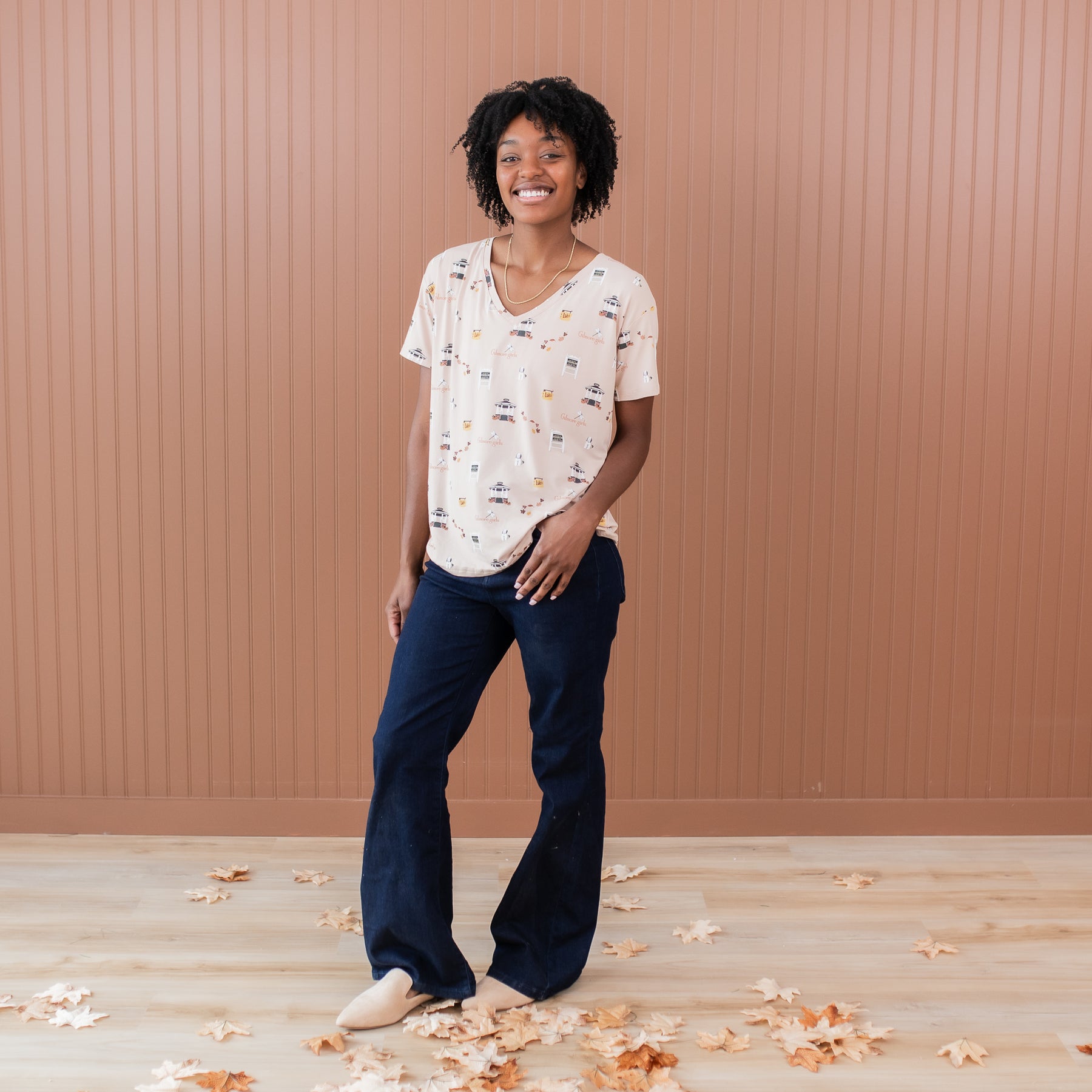 Female model standing in front of a light brown paneled wall wearing the Women's Relaxed Fit V-Neck in Gilmore Girls paired with dark wash jeans