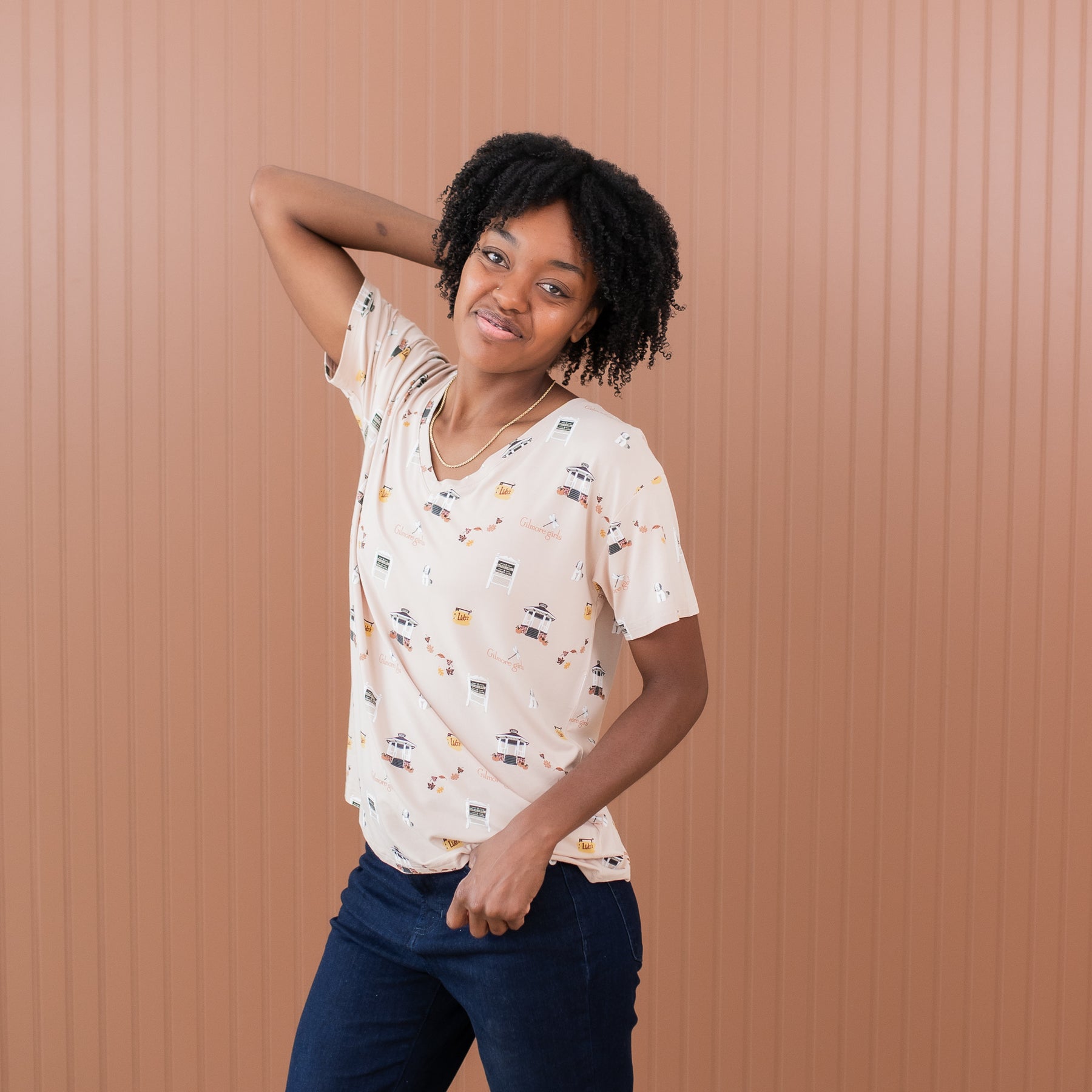 Female standing in front of a light brown paneled wall wearing the Women's Relaxed Fit V-Neck in Gilmore Girls with one arm behind her head