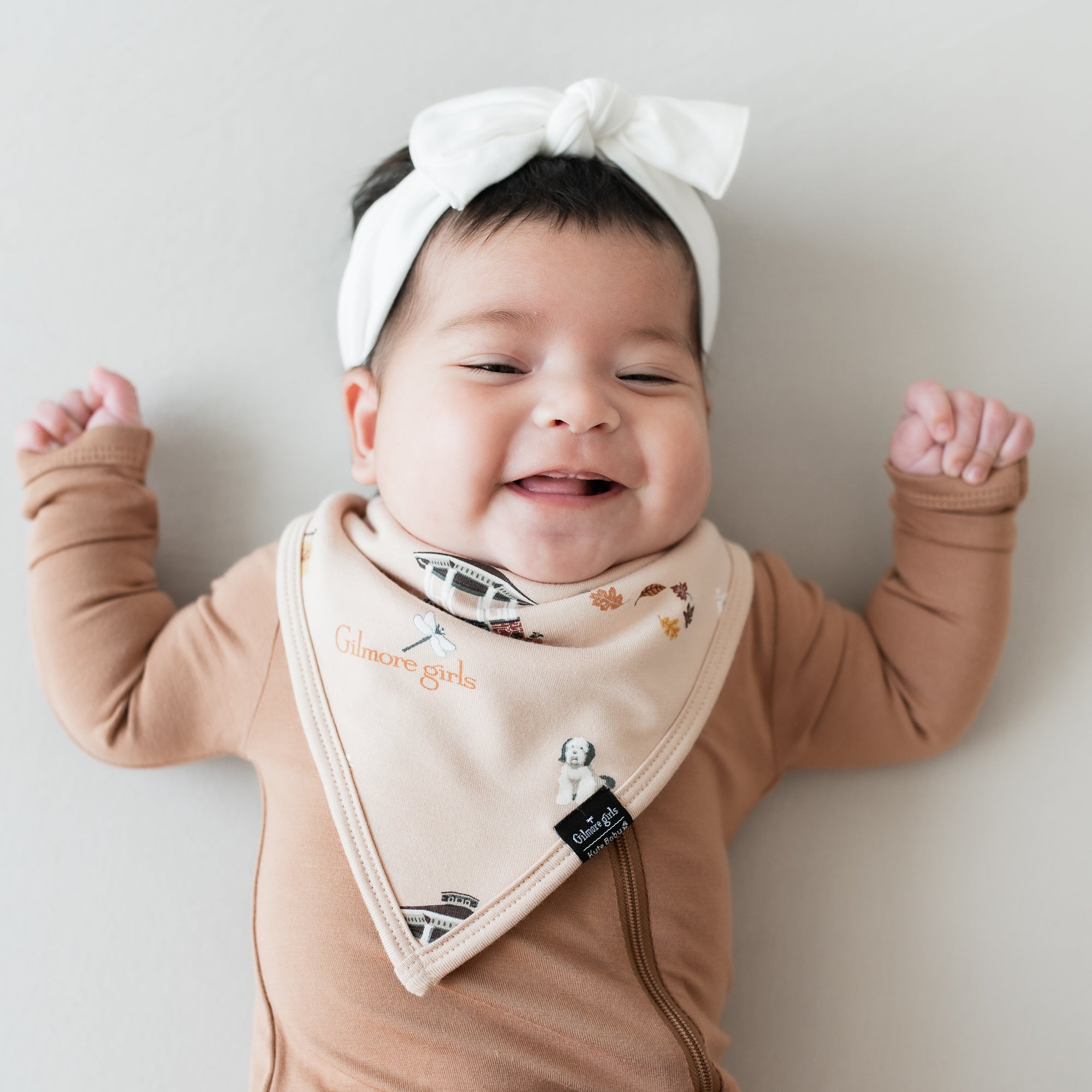 Smiling infant wearing the Bib in Gilmore Girls and Cloud Knotted Bow with a Latte zippered romper