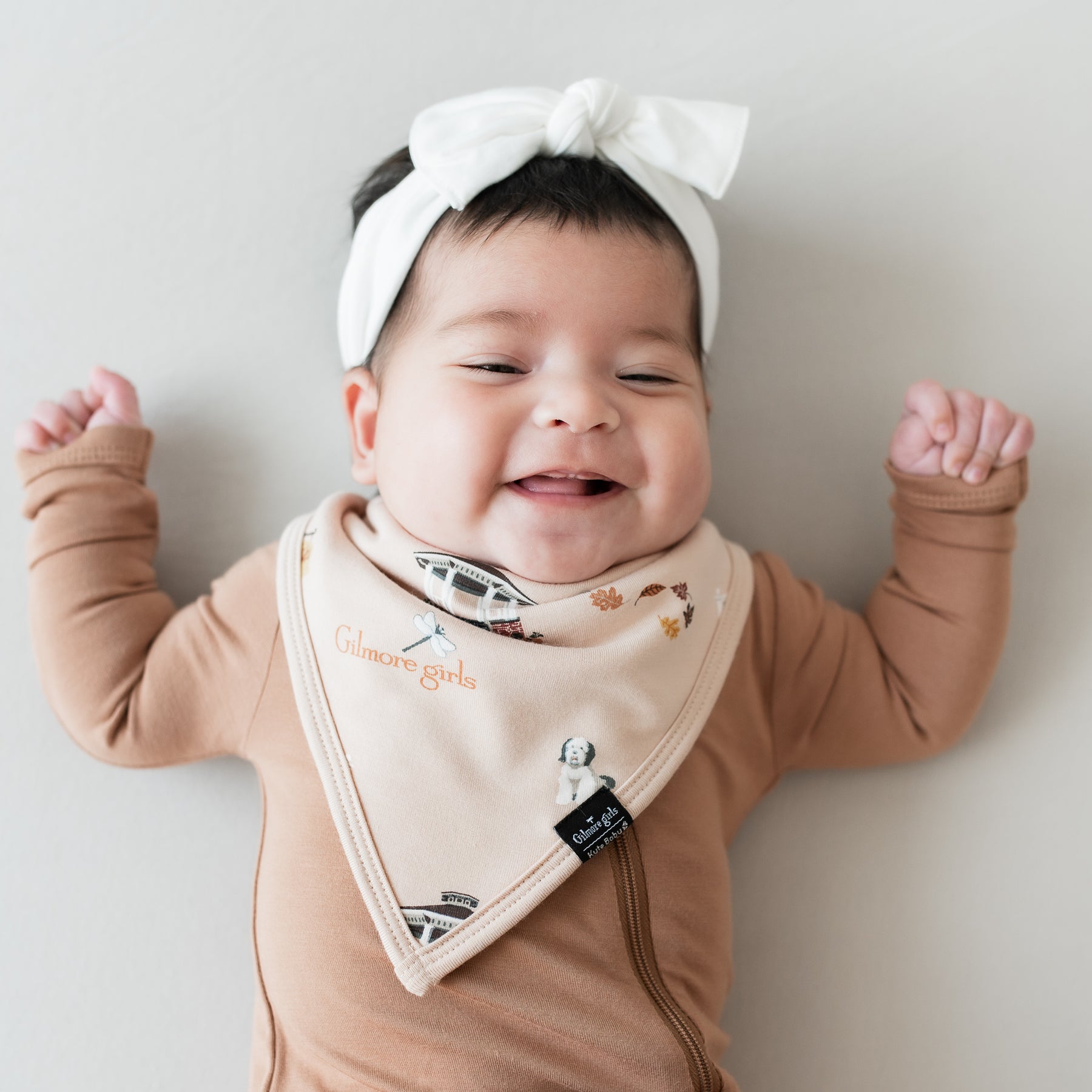 Smiling infant wearing the Bib in Gilmore Girls and Cloud Knotted Bow with a Latte zippered romper
