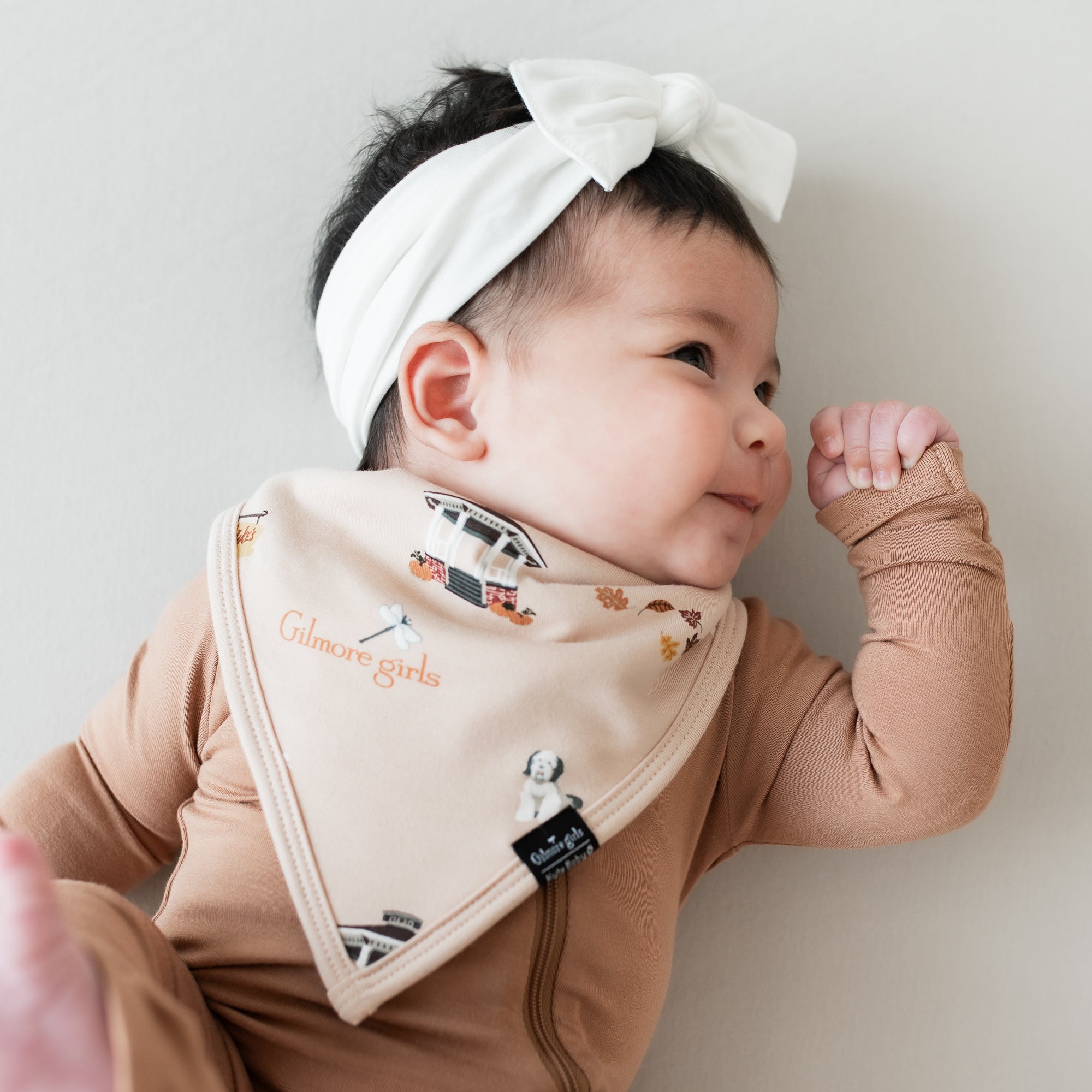 Infant looking to the side wearing the Bib in Gilmore Girls with a latte zippered romper and white bow