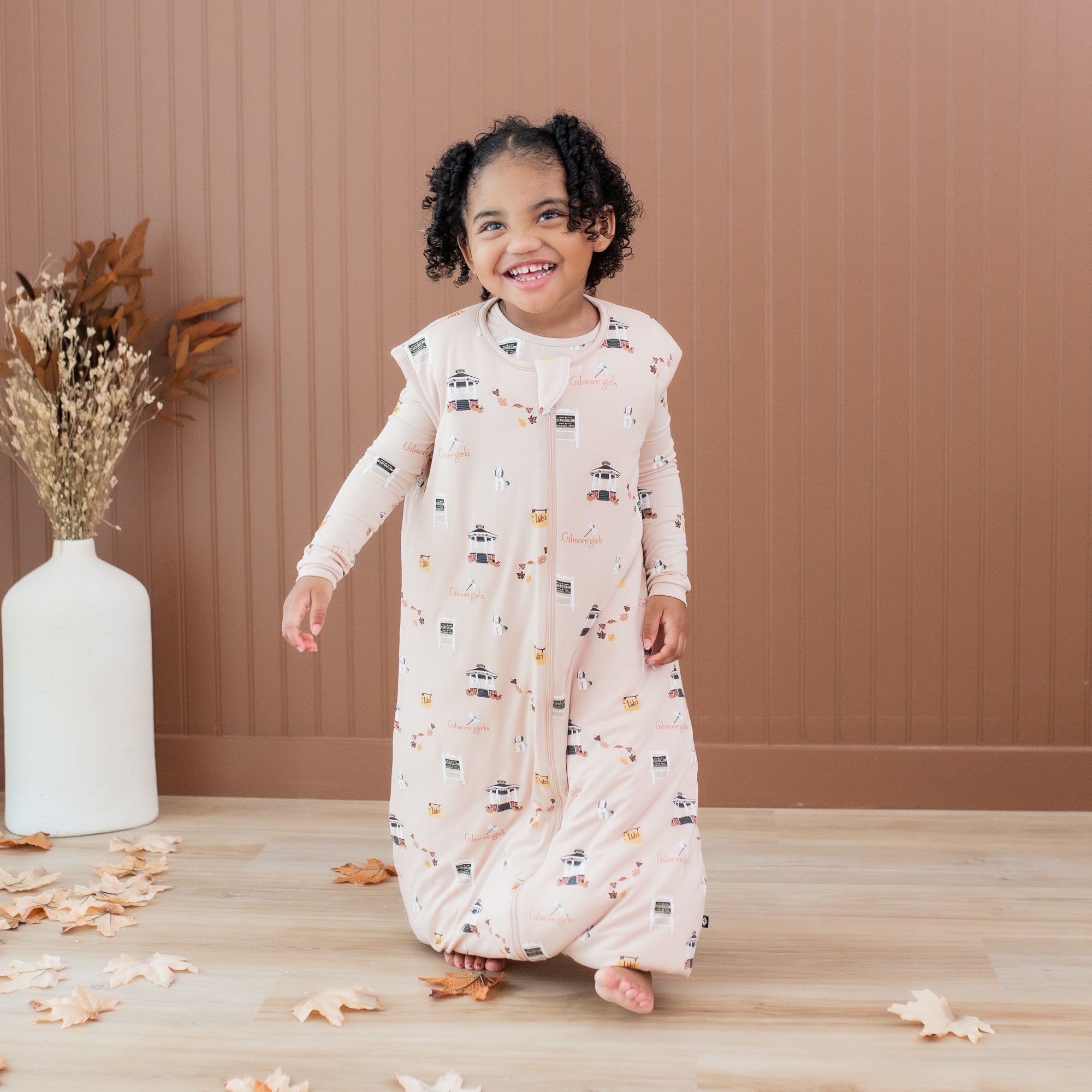 Smiling young girl wearing the Sleep Bag Walker in Gilmore Girls 1.0 walking in front of a light brown paneled wall with decorative leaves on the floor