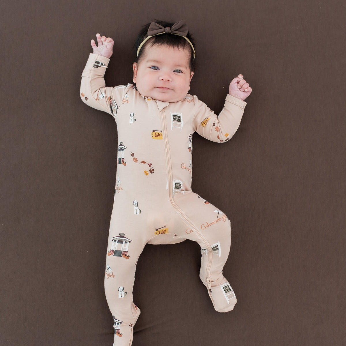 Infant girl laying on an Espresso crib sheet wearing the Zippered Footie in Gilmore Girls paired with a Tiny Nylon Bow in Espresso