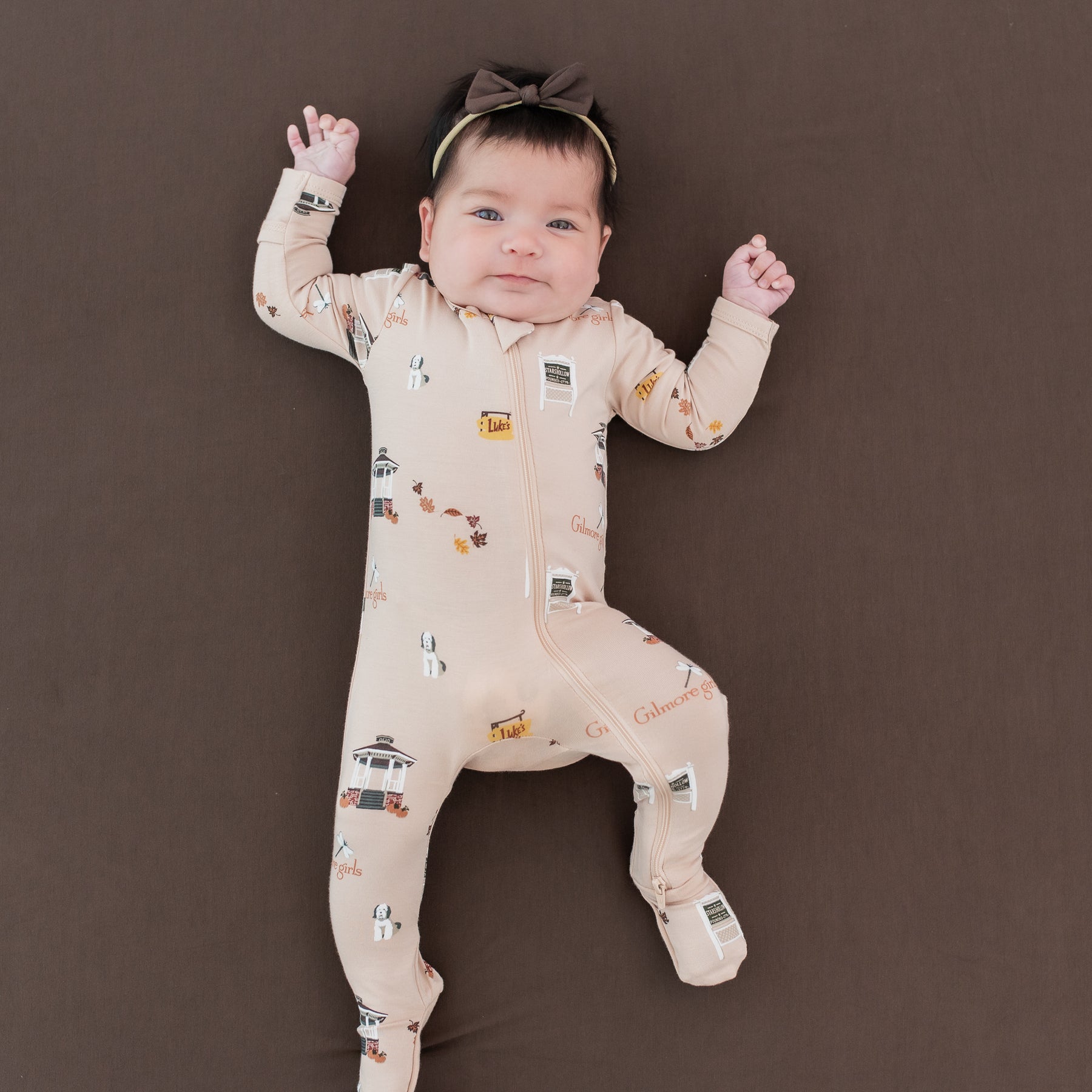 Infant girl laying on an Espresso crib sheet wearing the Zippered Footie in Gilmore Girls paired with a Tiny Nylon Bow in Espresso