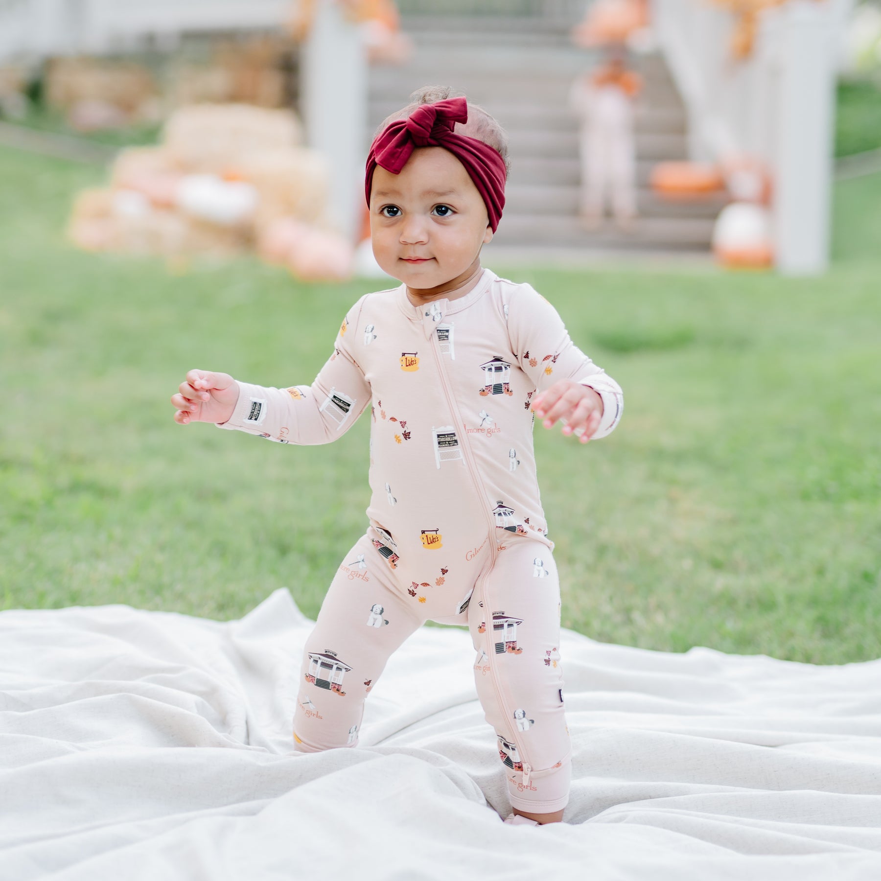 Young toddler in the grass standing on a cream colored blanket wearing the Zippered Romper in Gilmore Girls with a burgundy knotted bow headband