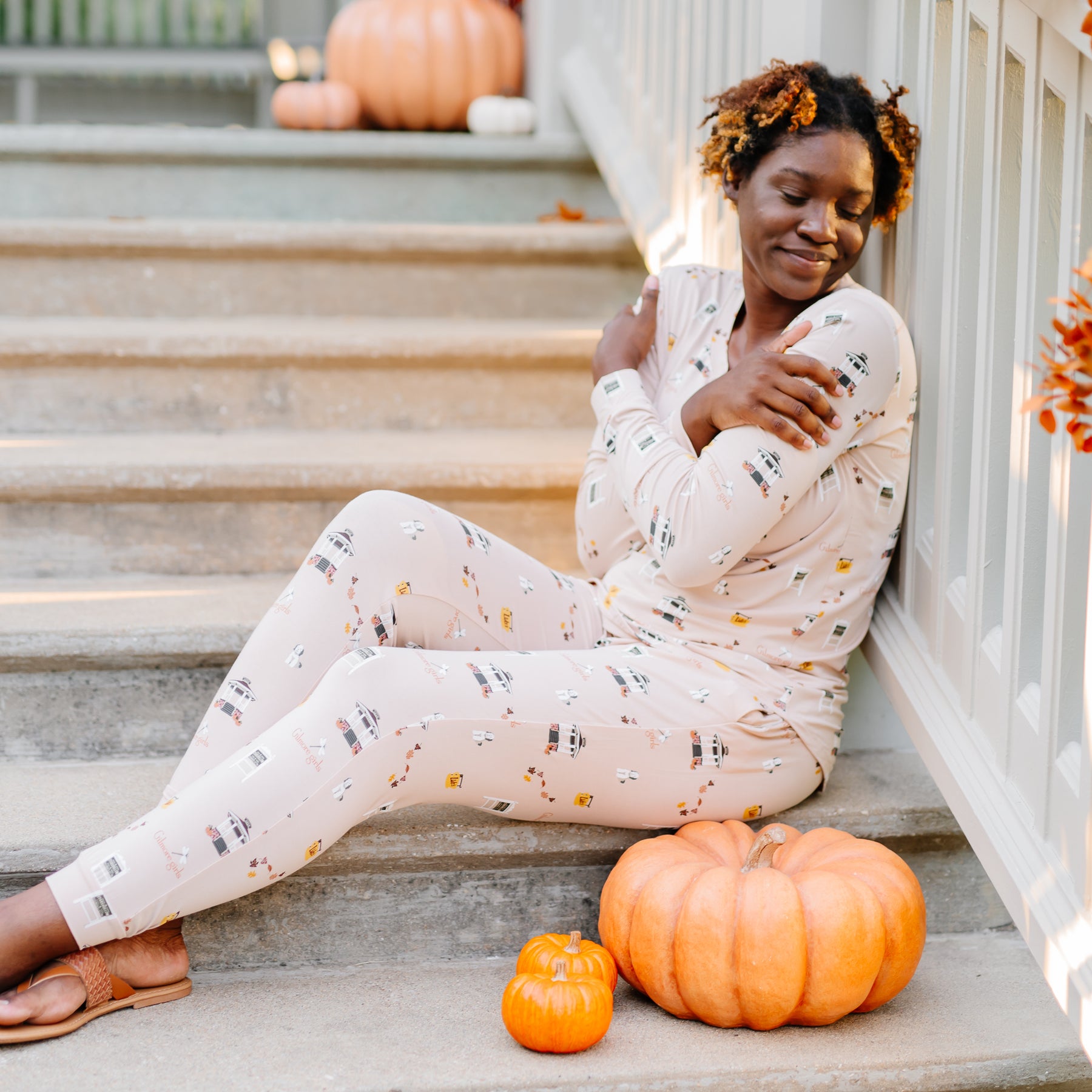 Female model sitting on stone steps leaning against a white railing wearing the Women's Jogger Pajama Set in Gilmore Girls with three pumpkins on the step in front of her