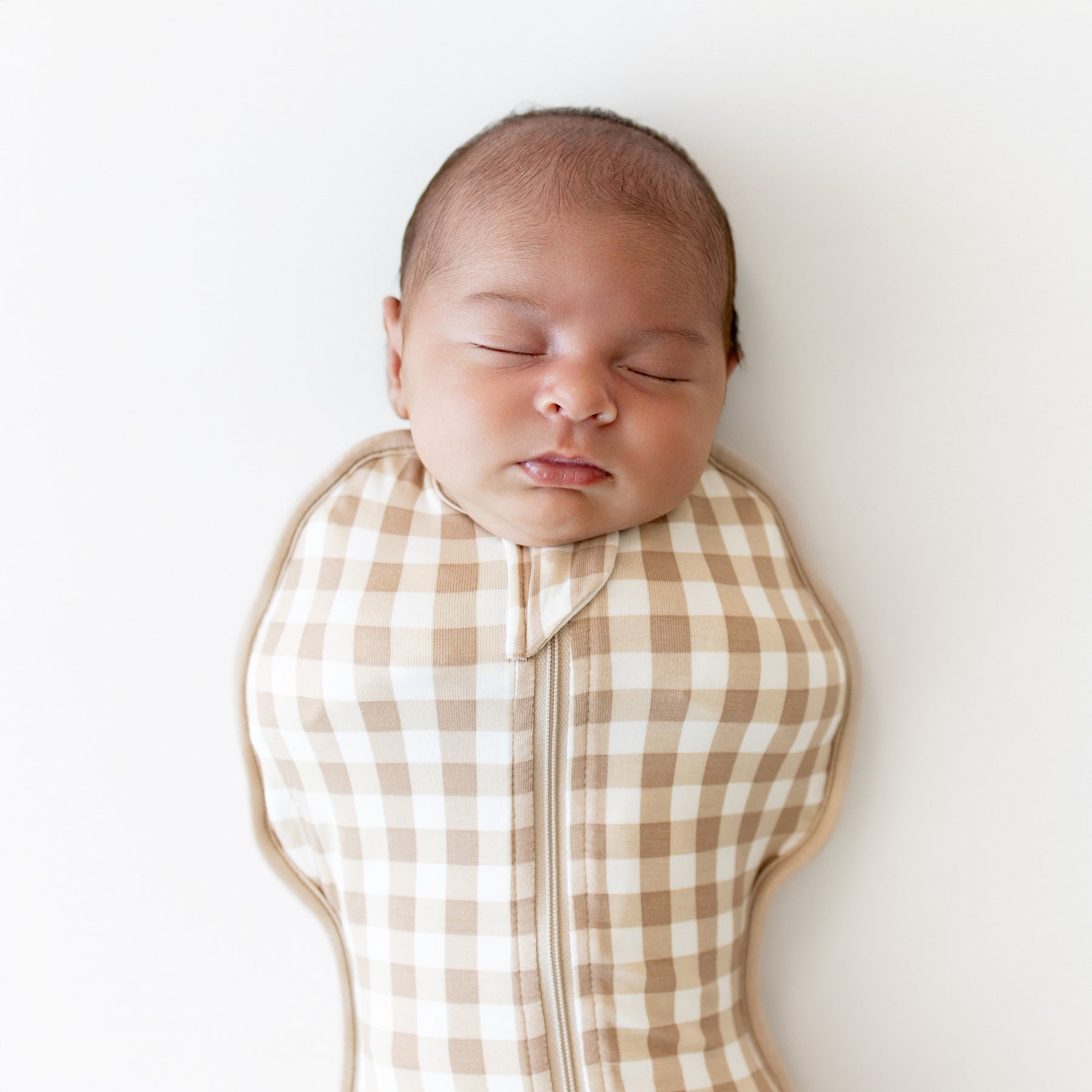 Newborn baby swaddled in a brown and white checkered swaddle bag on a white background