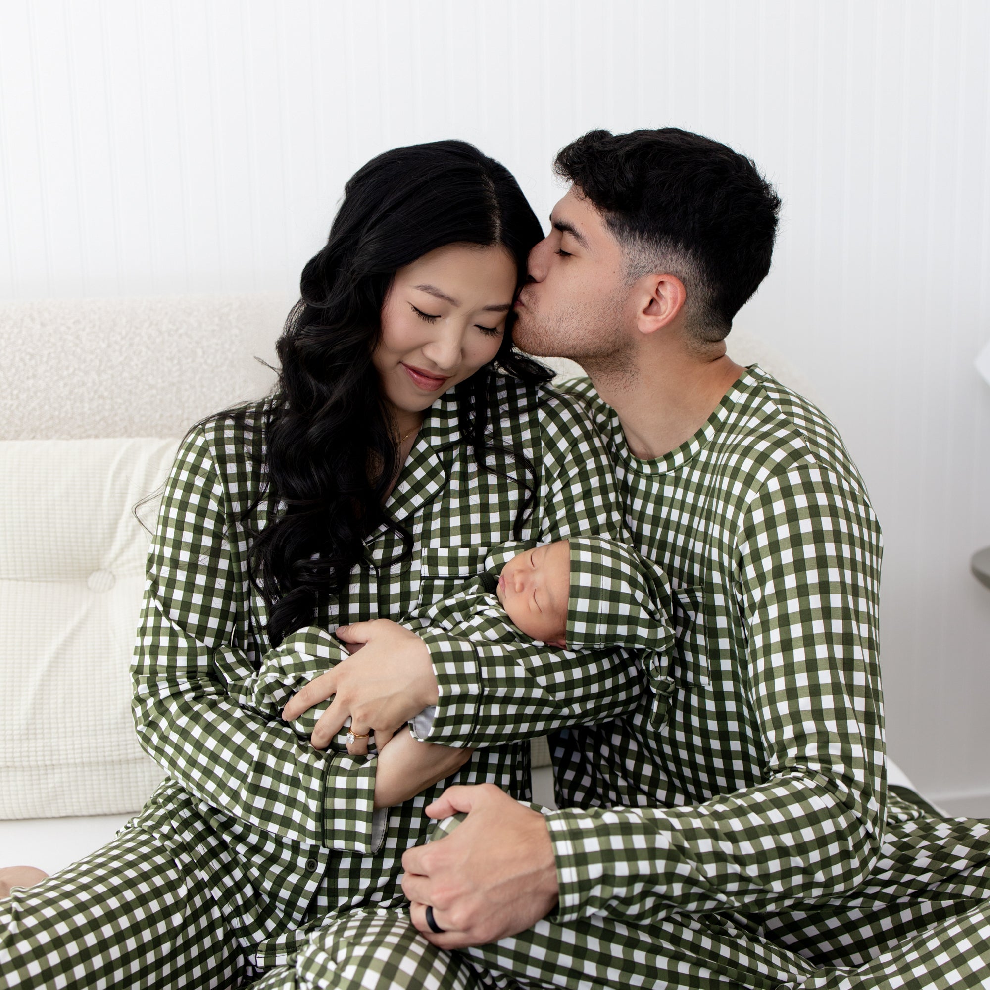 Couple in matching green checkered outfits holding a baby on a white couch.